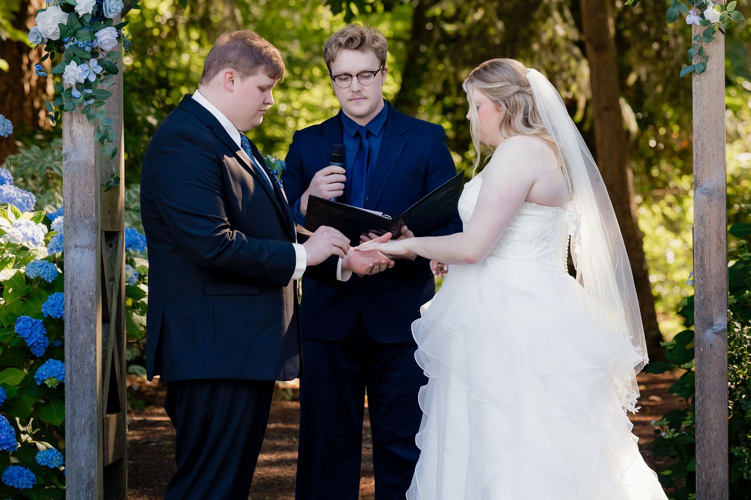 Outdoor wedding ceremony with bride and groom exchanging rings before an officiant, framed by blue floral arch in a wooded setting.