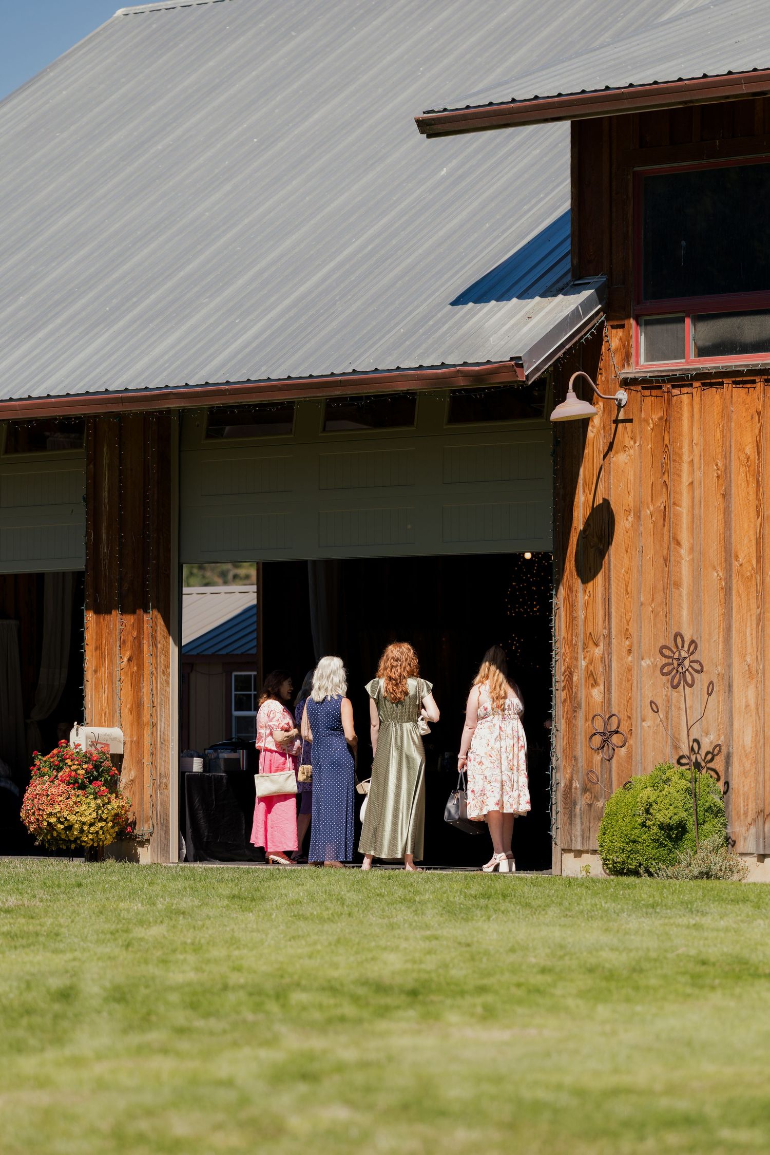 Four women in elegant dresses gather at the entrance of a rustic wooden barn wedding venue on a sunny day.