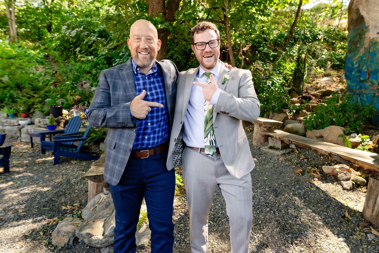 Two smiling men in suits give thumbs up together at an outdoor wedding venue surrounded by greenery.