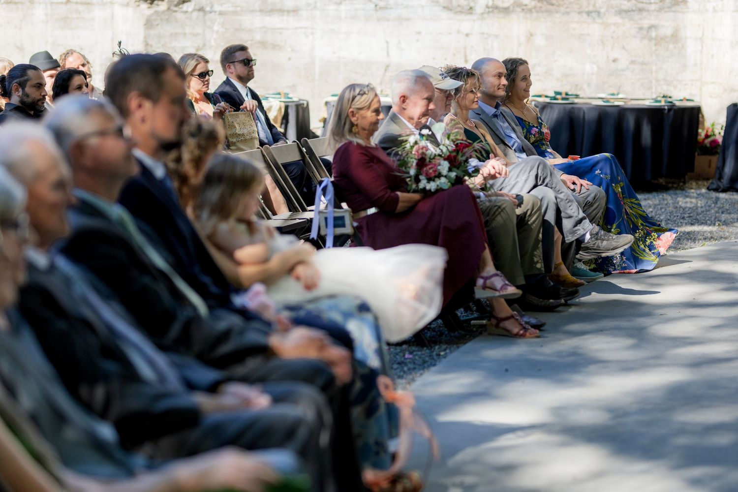 Guests seated outdoors at a ceremony, dressed in formal attire, attentively watching the event unfold.