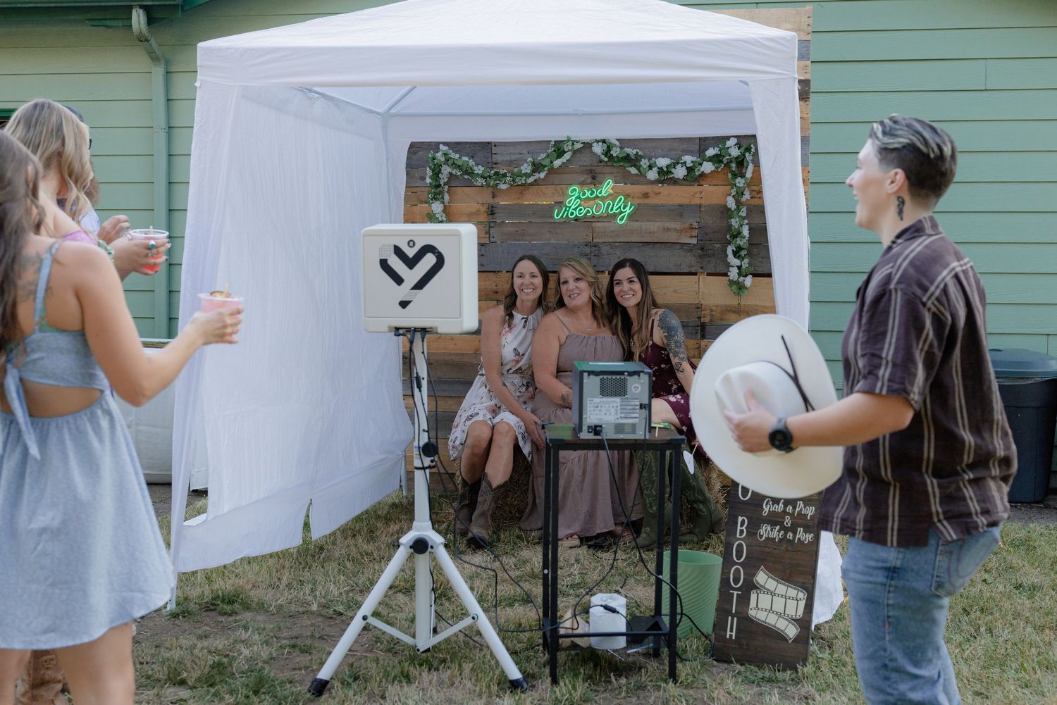 Guests pose at outdoor wedding photo booth with neon sign under white canopy tent.