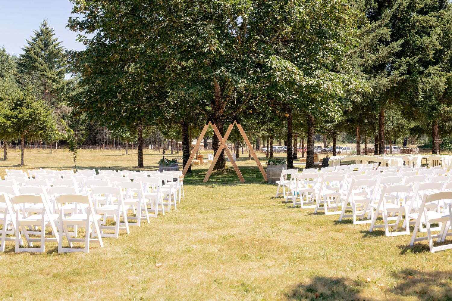 Outdoor wedding ceremony setup with white folding chairs, wooden geometric arch, and tall trees in background on sunny day.