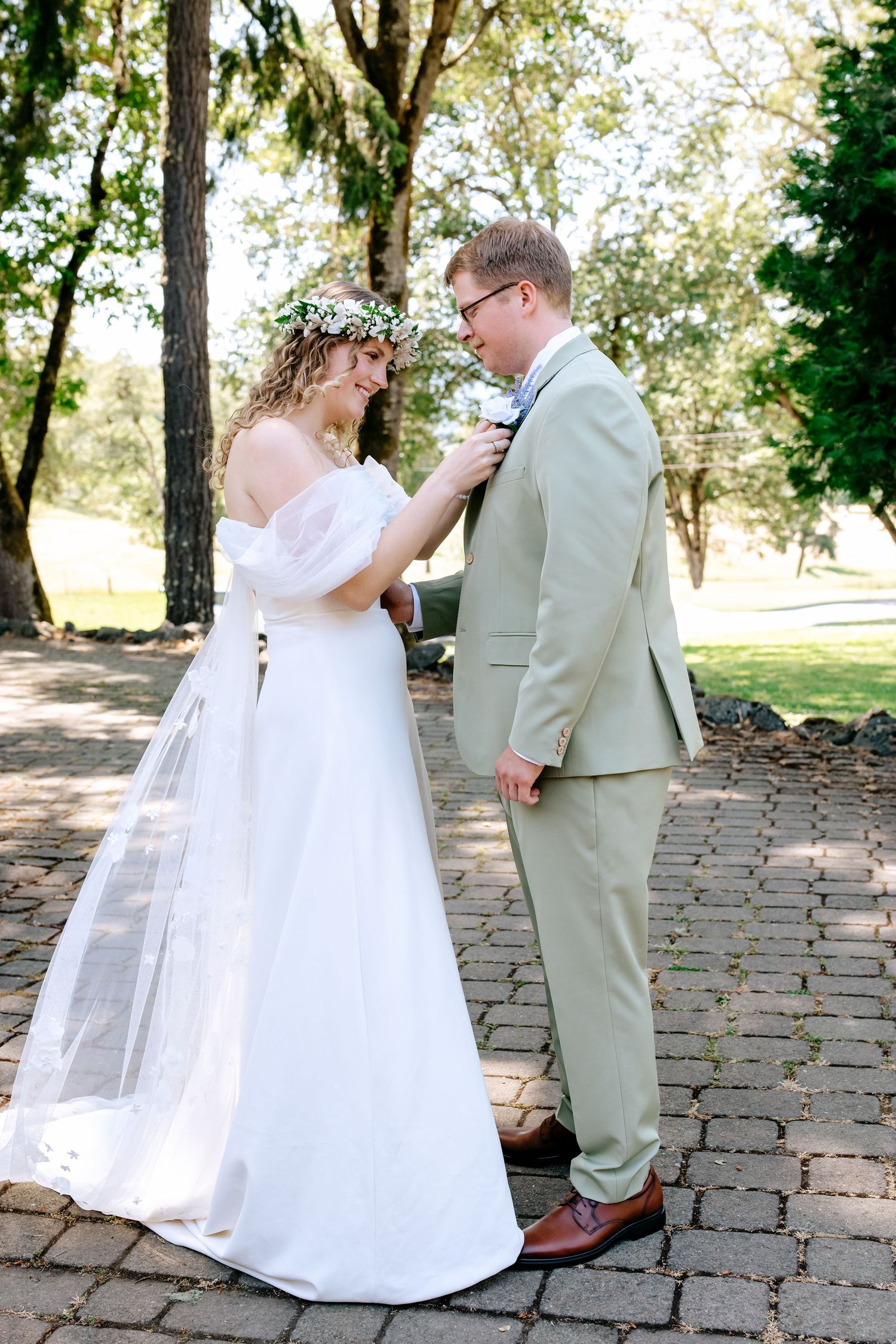 Bride adjusting groom's boutonniere on his sage suit while wearing a floral crown on their wedding day outdoors.
