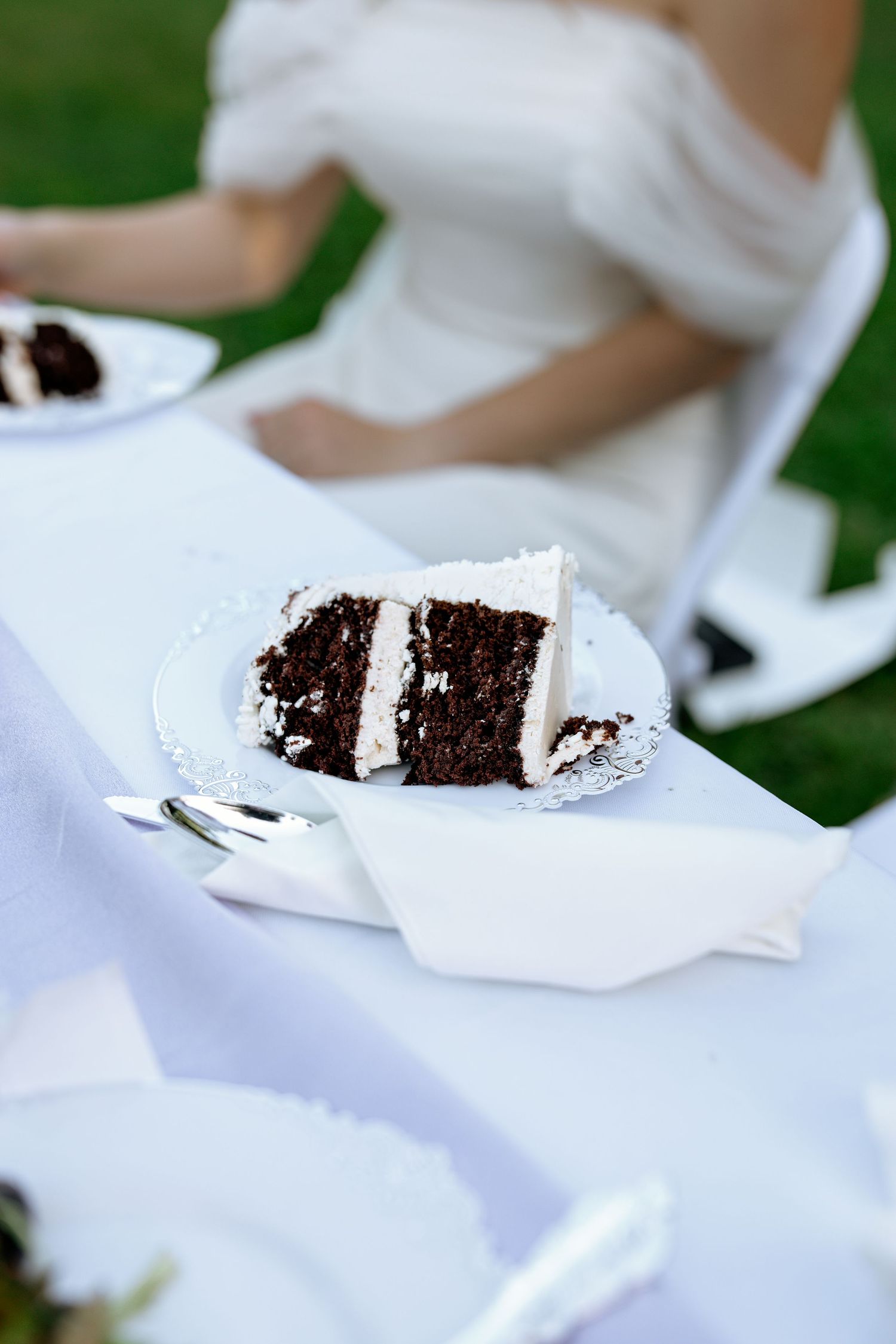 Slice of chocolate layer cake with white frosting served on white plate at outdoor wedding reception.