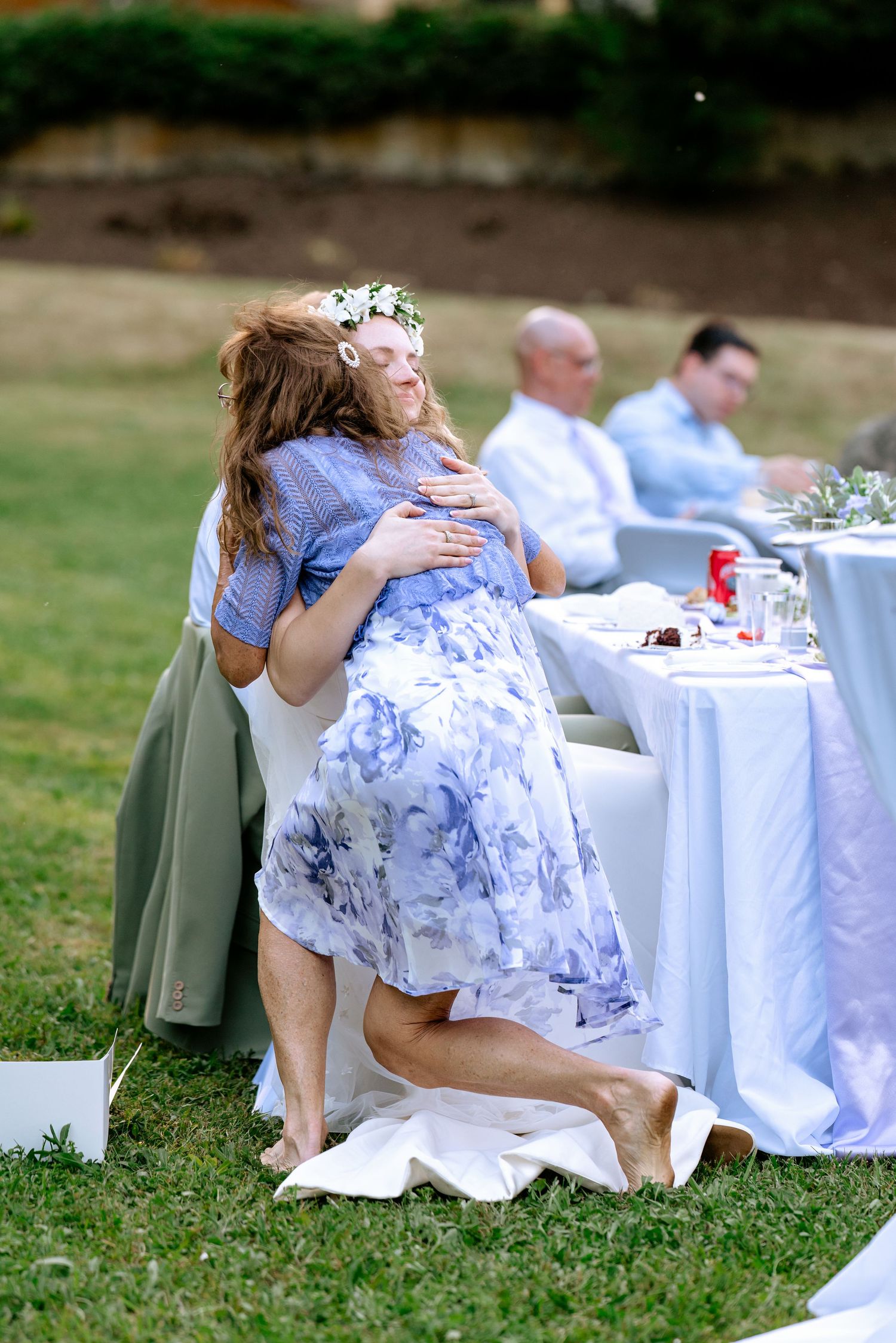 Bride in floral crown shares a warm embrace with a guest kneeling on the grass at reception.
