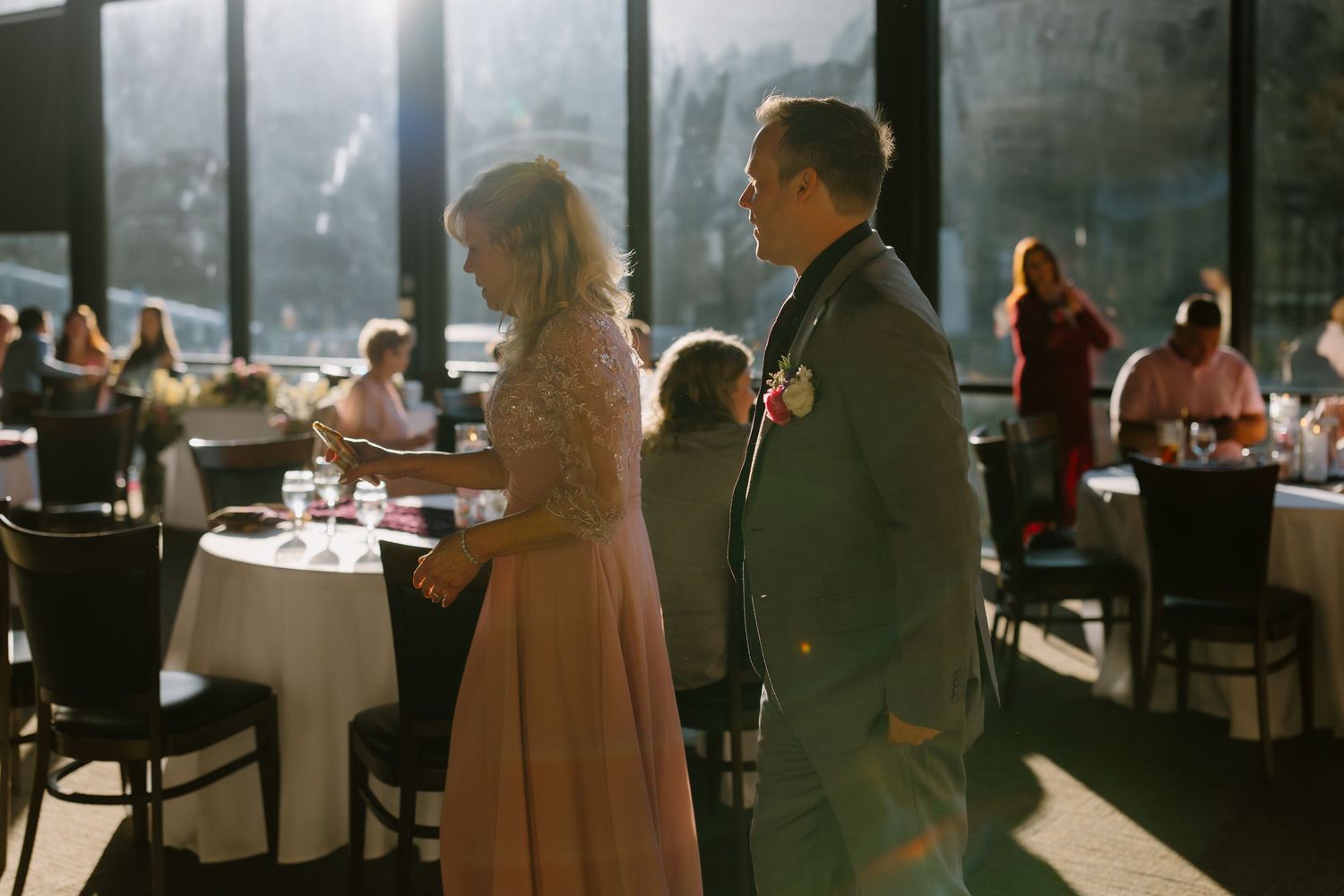 Couple in elegant attire holding hands at a sunlit wedding reception venue with guests seated at tables.