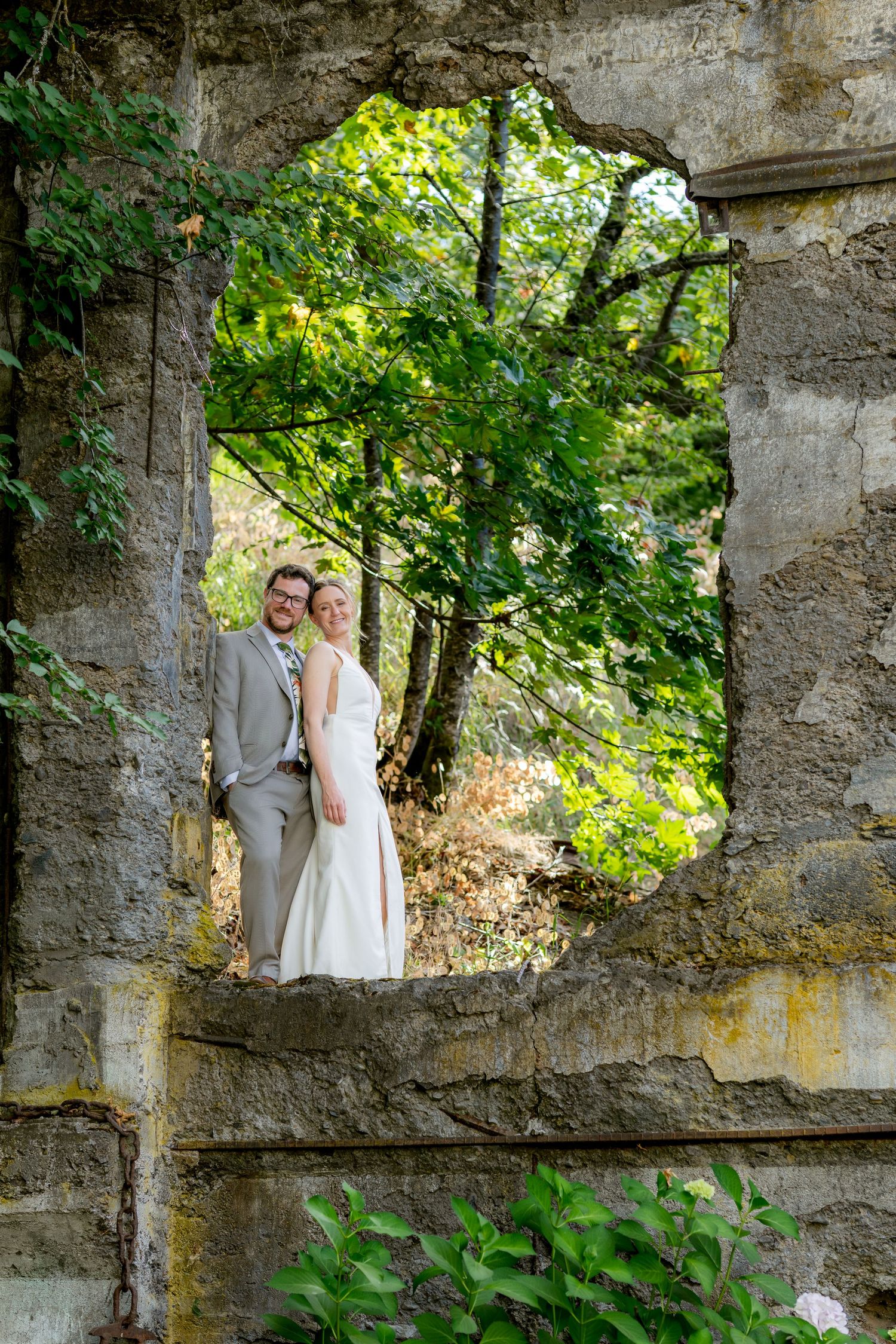 Bride and groom posing in a rustic stone window frame surrounded by lush green trees in an outdoor wedding photo.