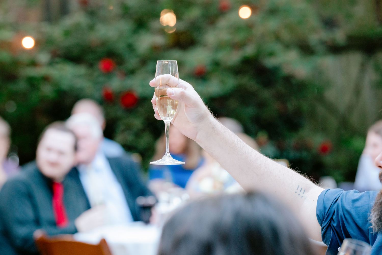 A hand raises a champagne flute in a toast at an outdoor evening wedding reception with guests seated in background.