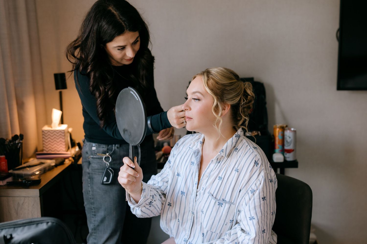 Makeup artist applying makeup to a seated bride holding a hand mirror during bridal preparation.