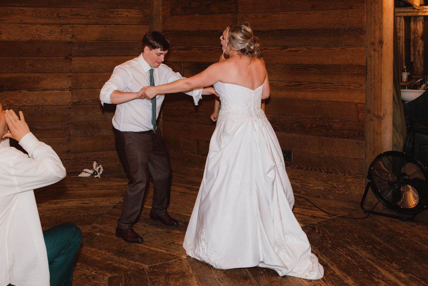 Bride in white ball gown dances with young man in white shirt inside rustic barn venue.