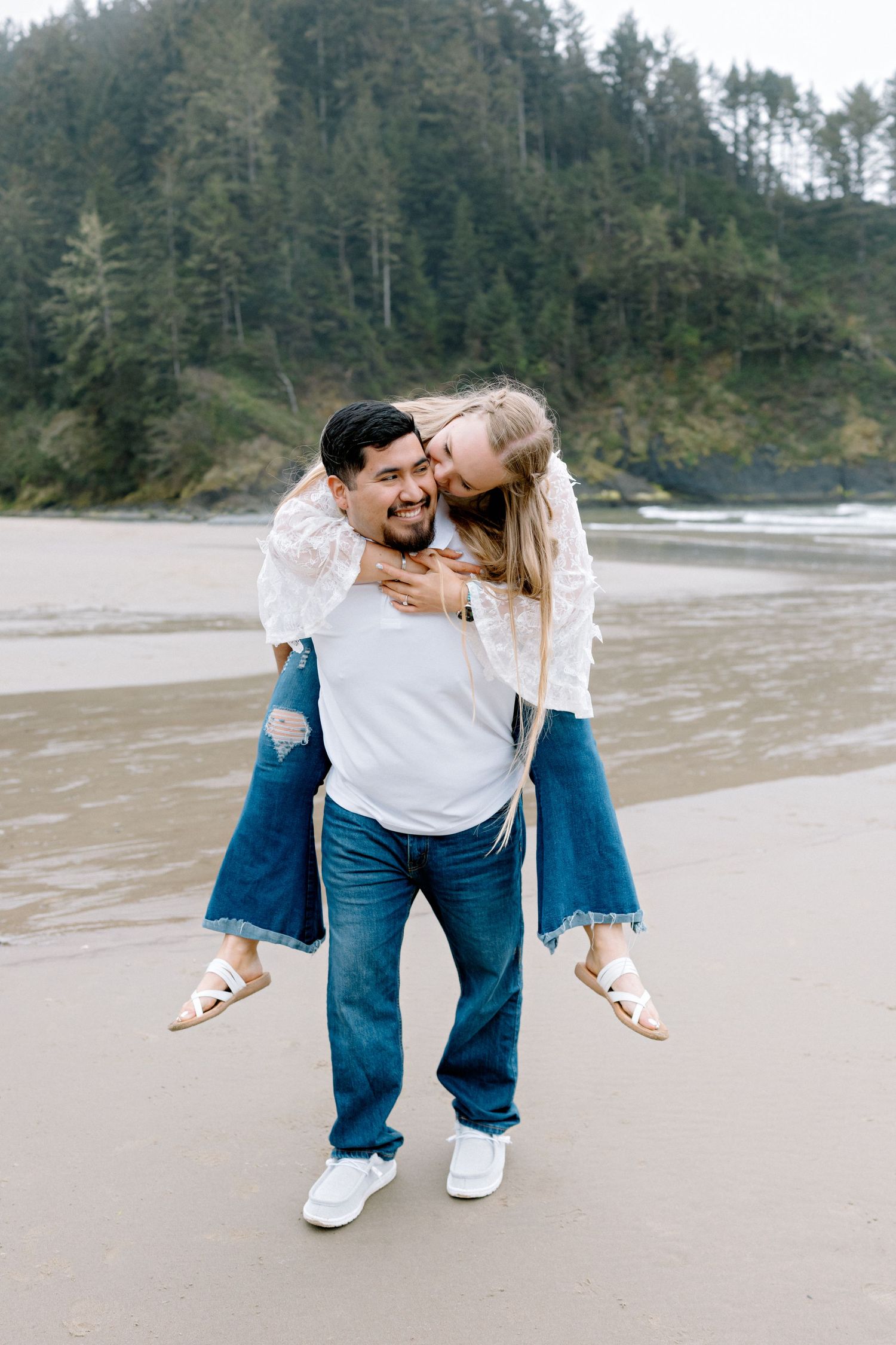 Man giving woman a piggyback ride on a beach, both smiling, with evergreen forest in the background.