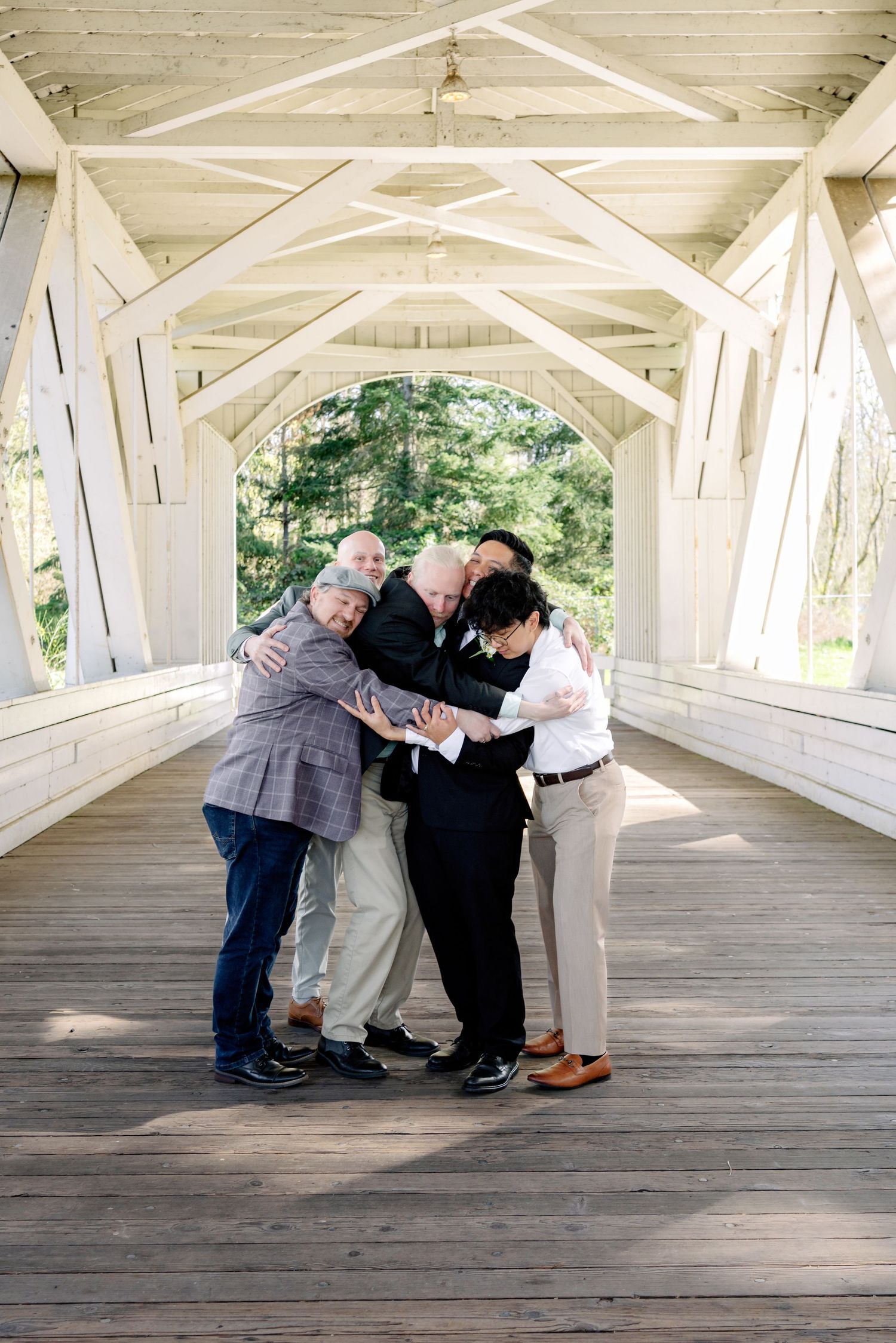 Newlyweds and family share joyful group hug inside white covered wooden bridge after wedding.