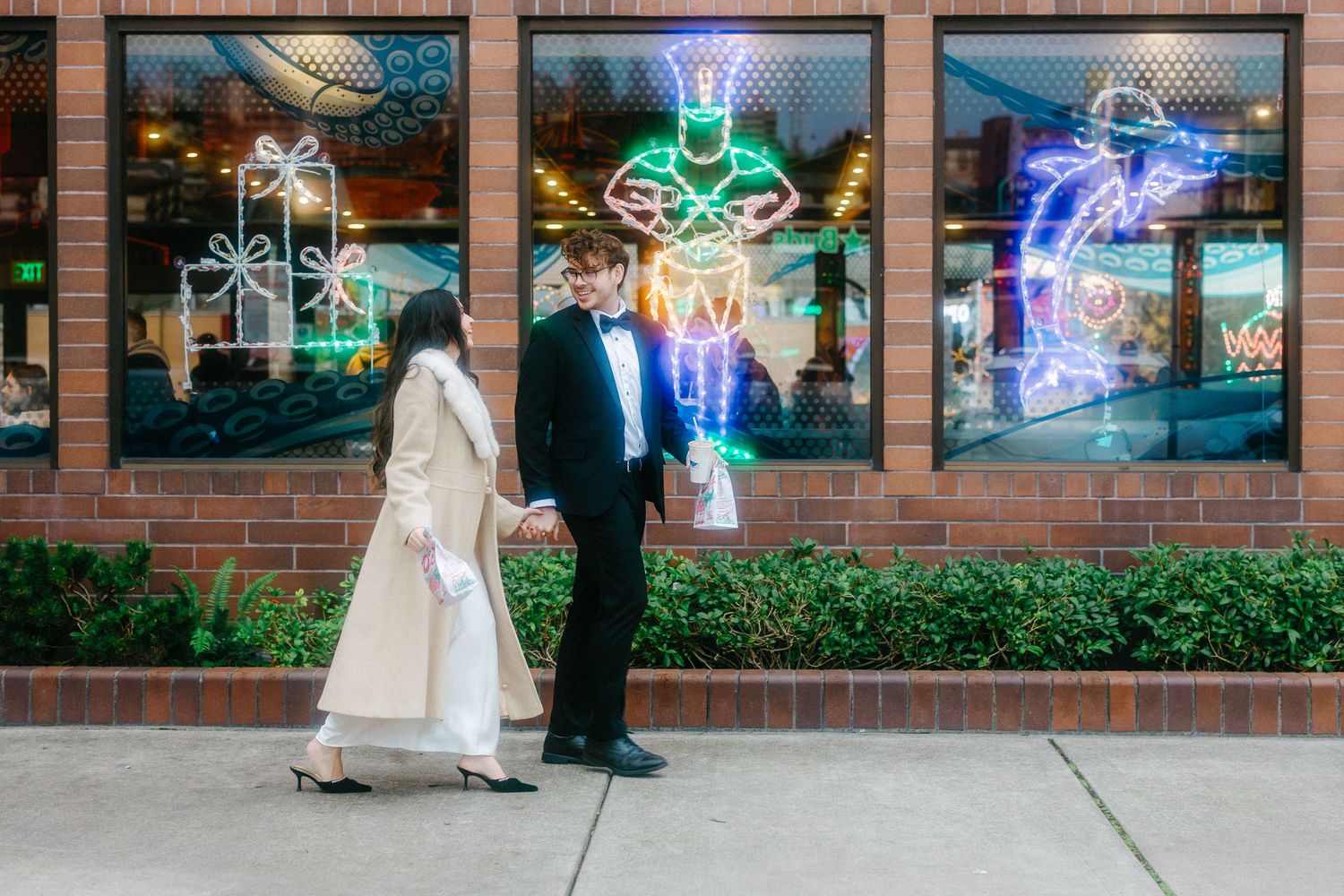 Two people stand on a sidewalk in front of colorful neon signs in storefront windows at dusk.