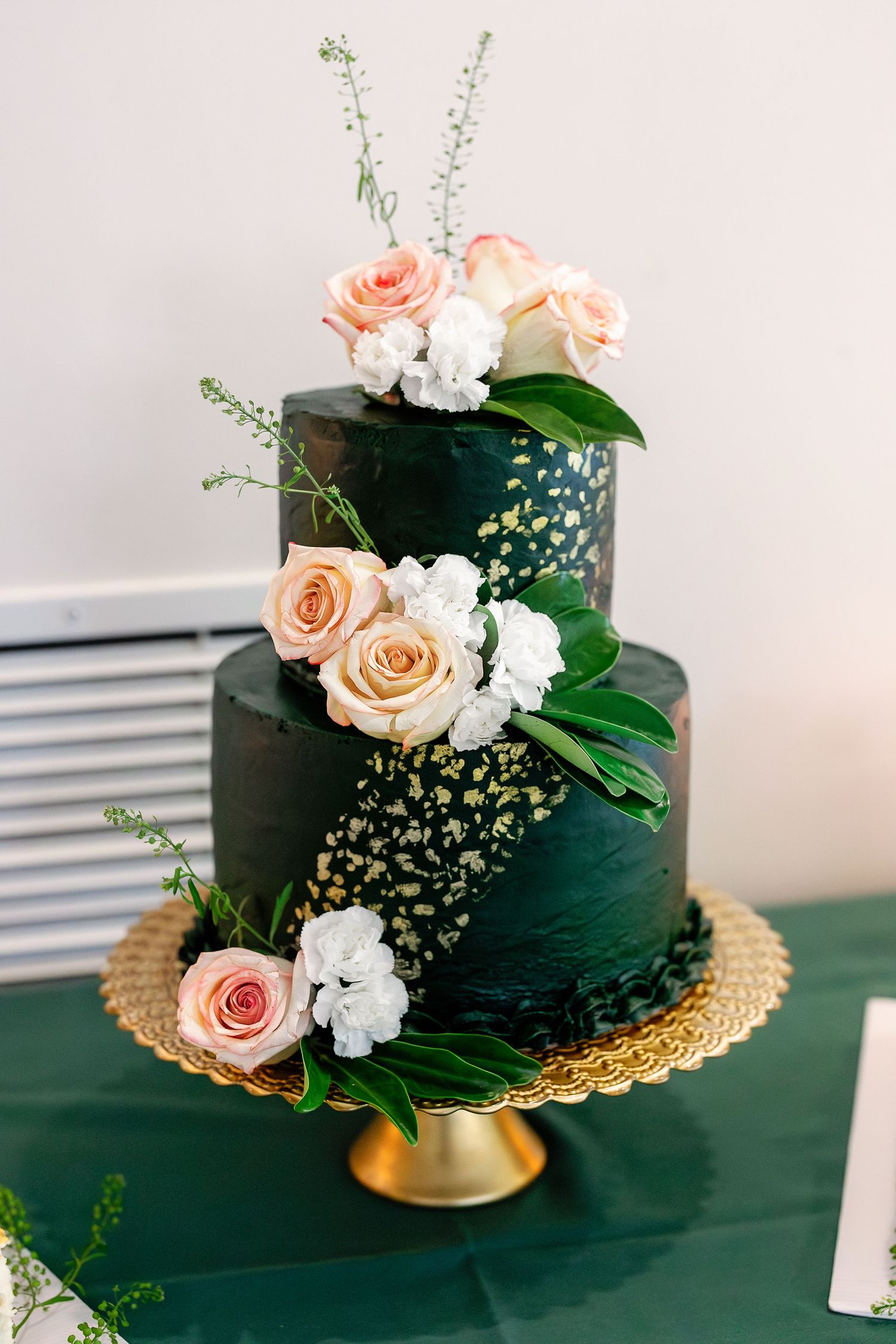 Elegant two-tier dark green wedding cake decorated with peach roses and white flowers on a gold stand.