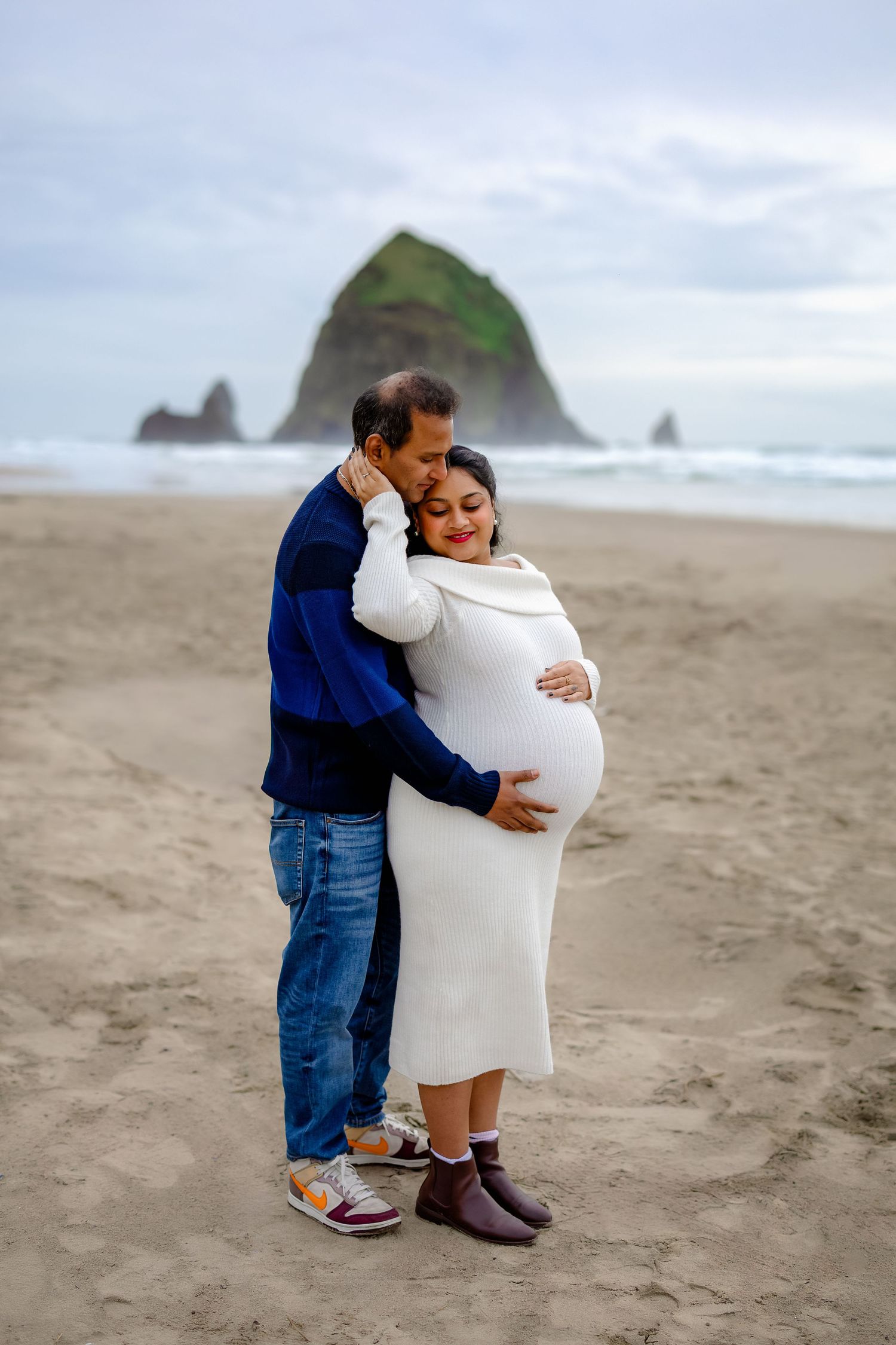 Two people embrace while standing on a sandy beach with coastal rock formations in the background.