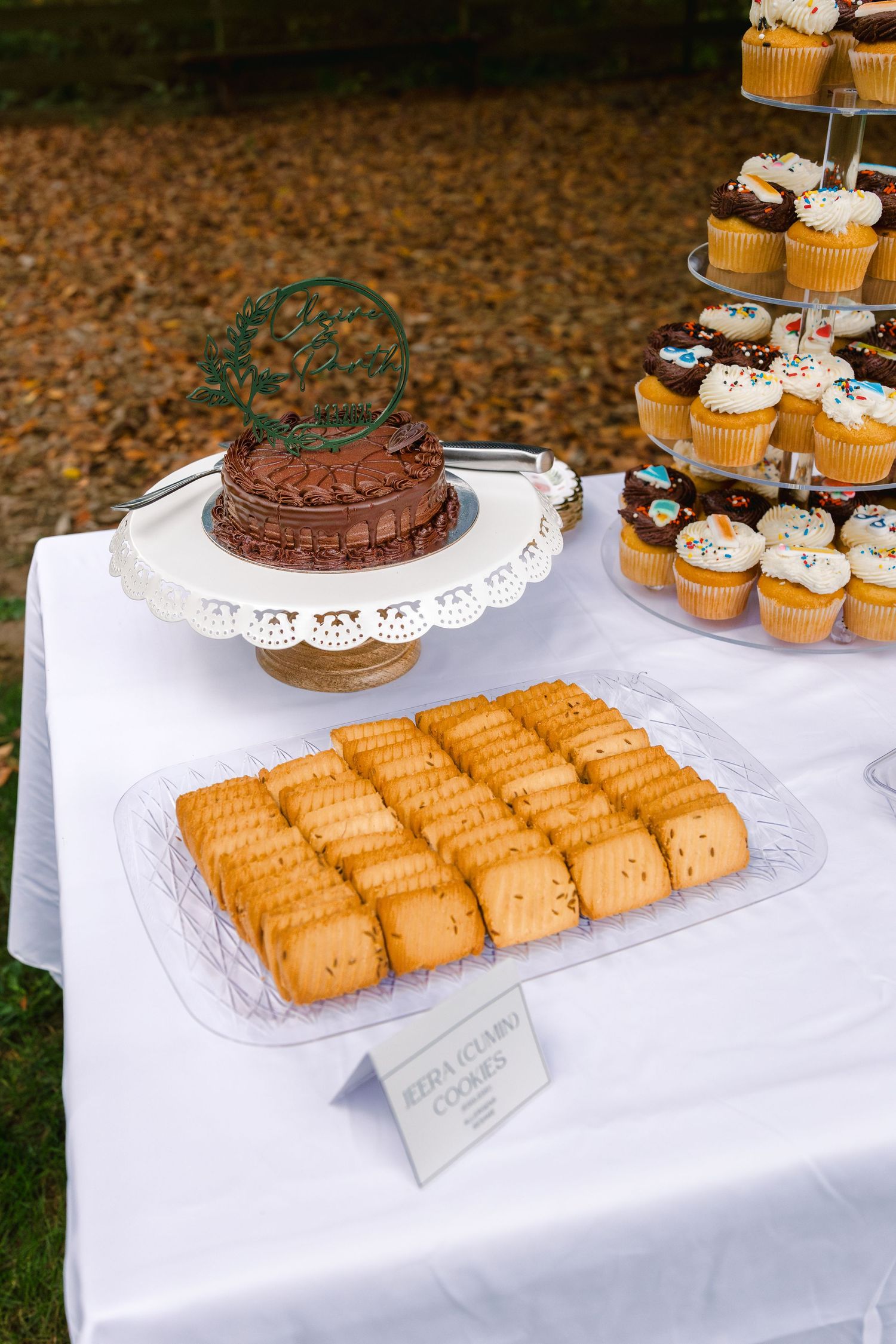 A dessert table displays chocolate cake, cupcakes and cookies on white tablecloths at an outdoor event.