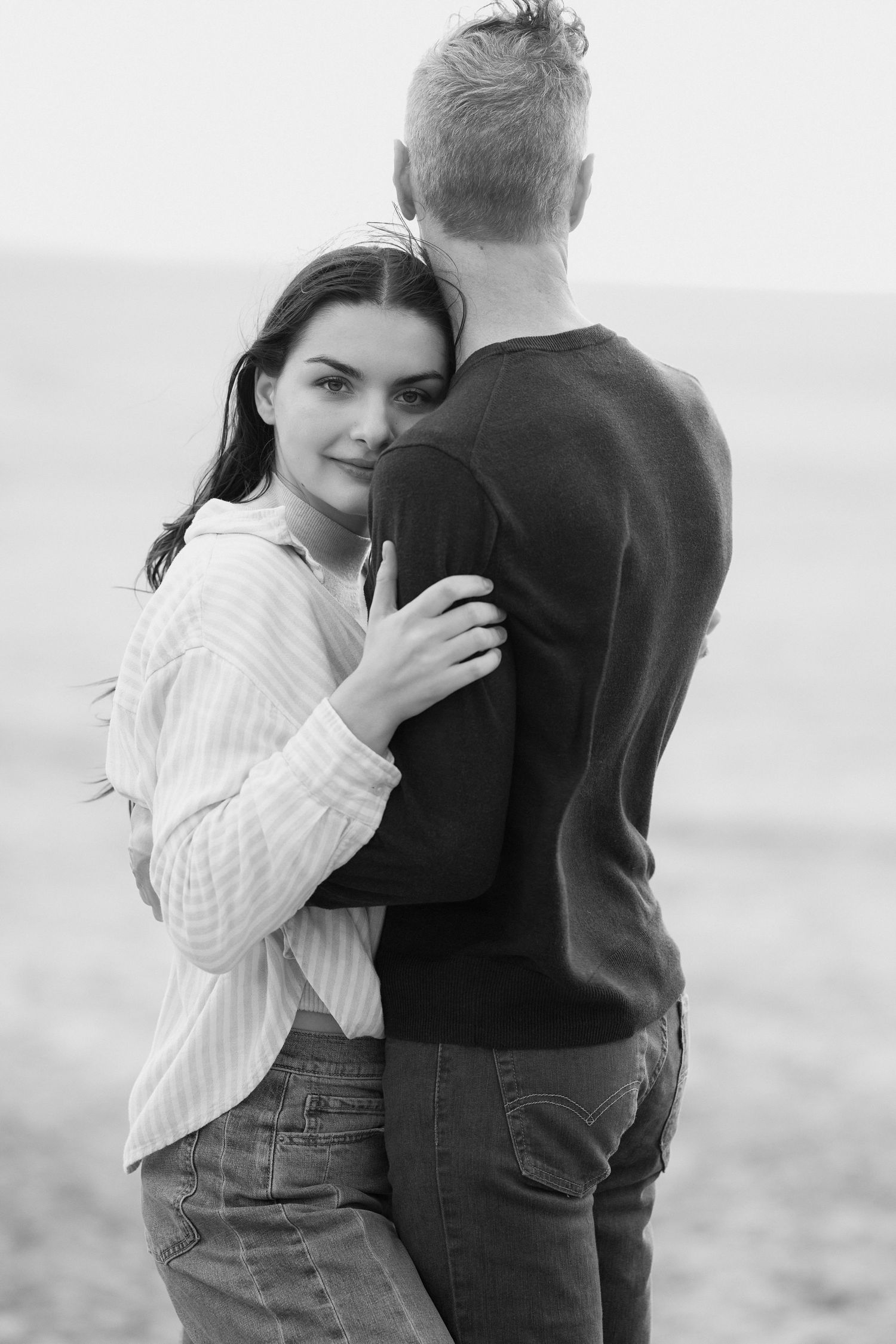 A romantic black and white portrait of a couple embracing on a beach in casual sweaters and jeans.