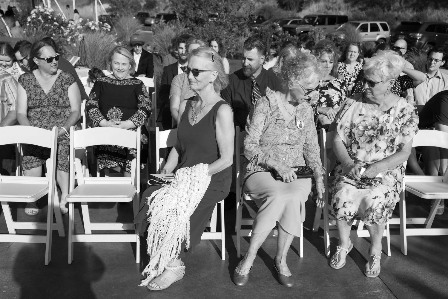 Black and white photo of guests seated in rows at an outdoor event with cars visible in background.