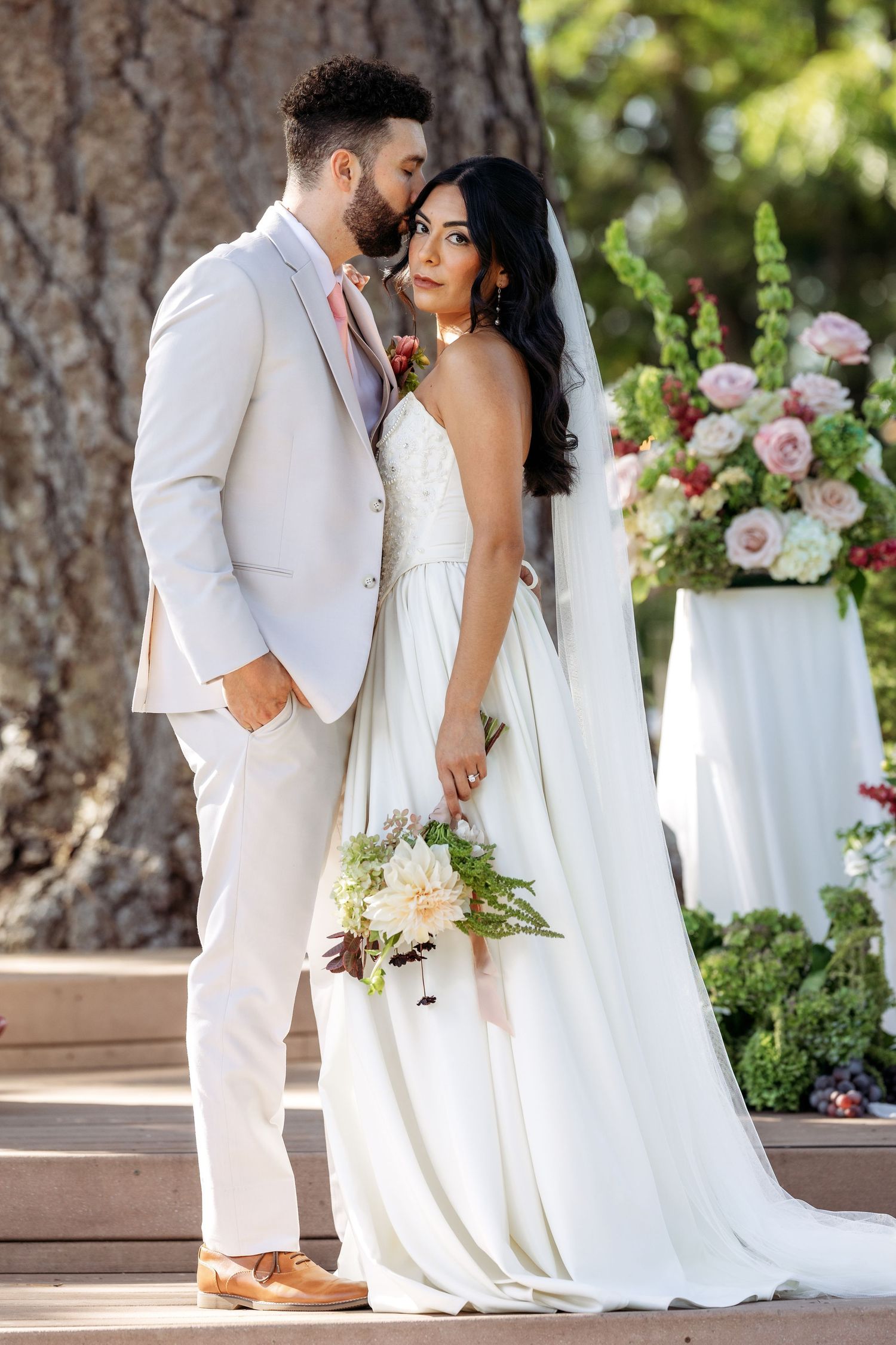 A couple in white wedding attire share an intimate moment outdoors with pink floral arrangements.