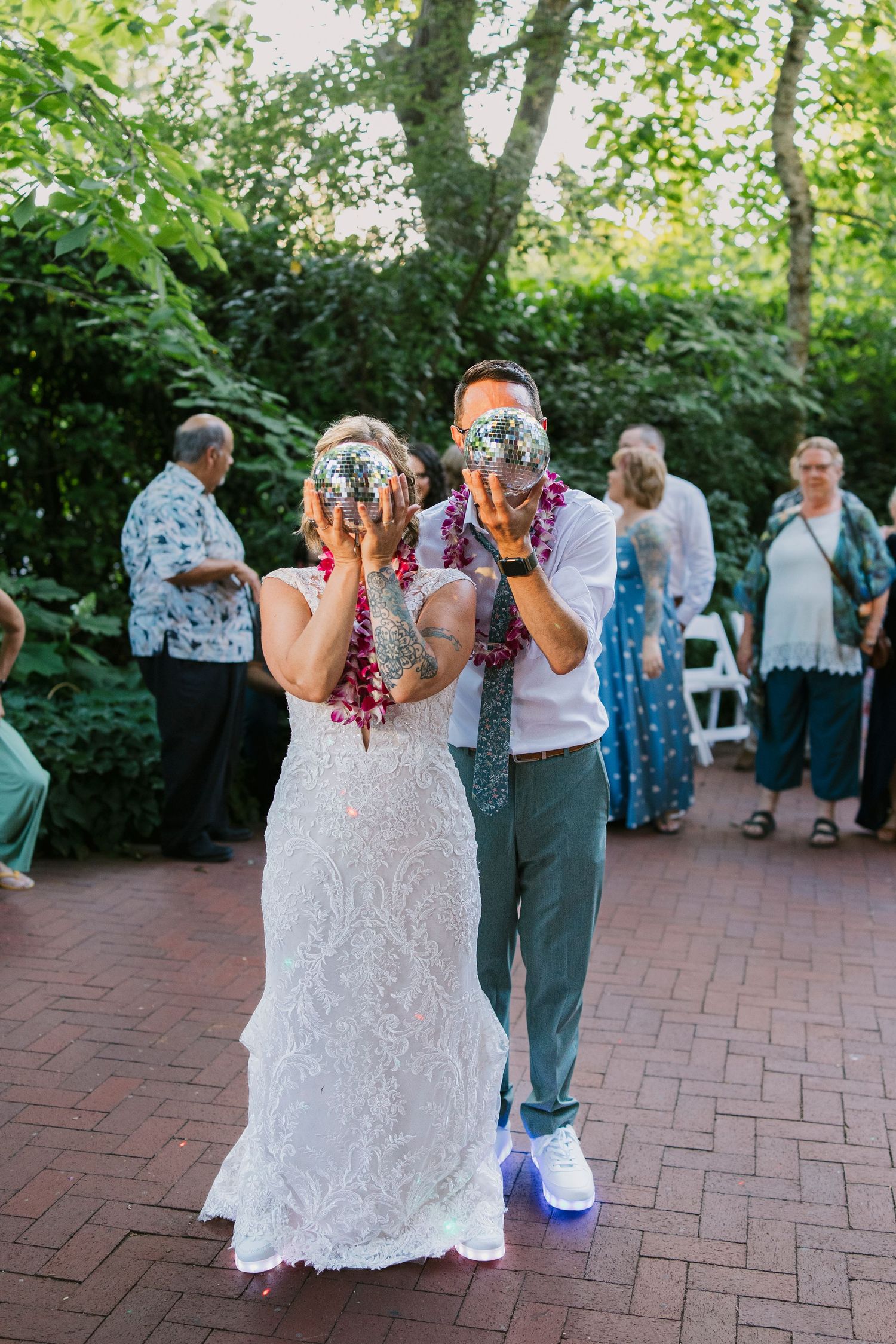 A couple shares their first drink at an outdoor garden wedding reception on a brick patio with guests in the background.