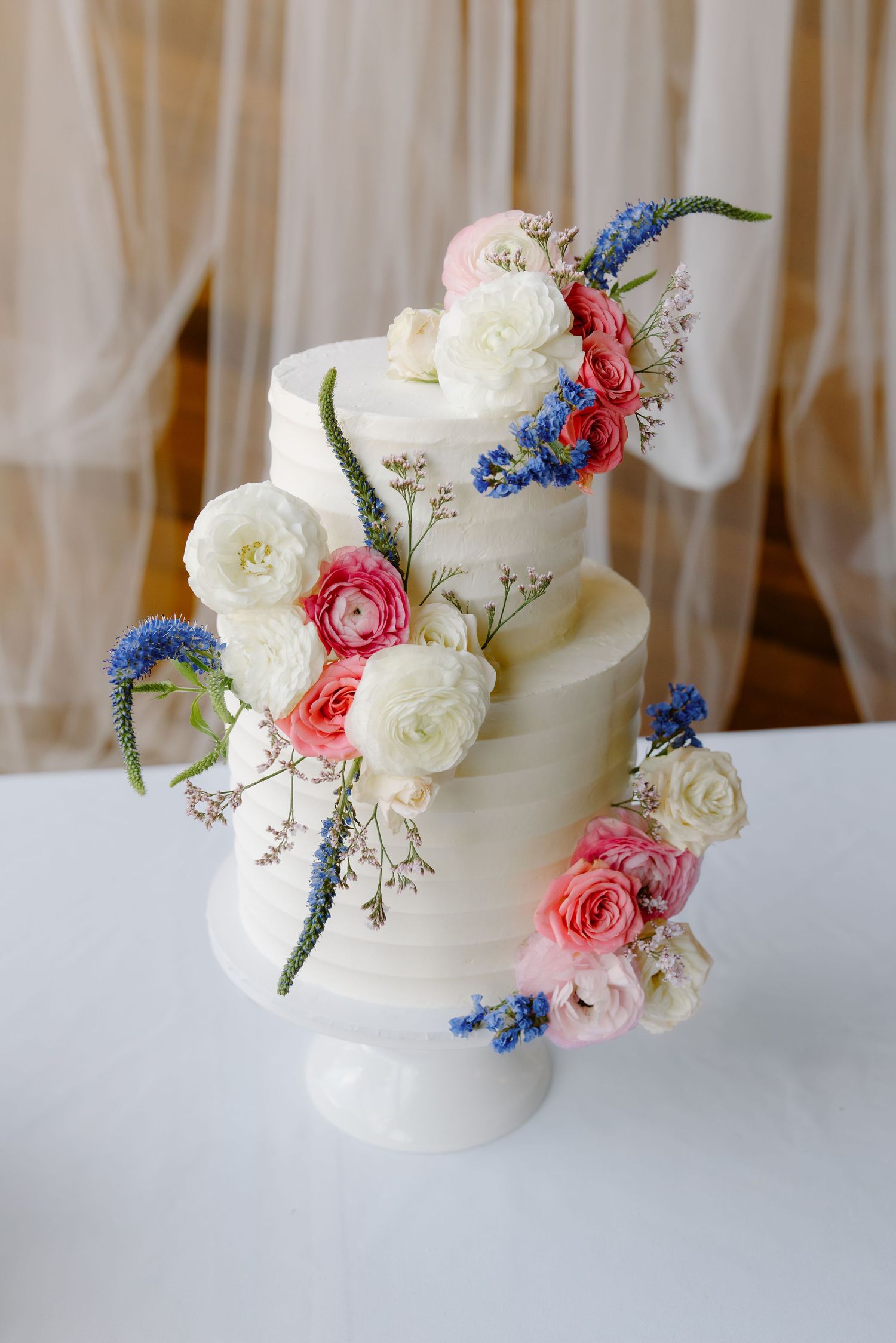 Elegant two-tier white wedding cake adorned with pink, white, and blue fresh flowers cascading down its layers.