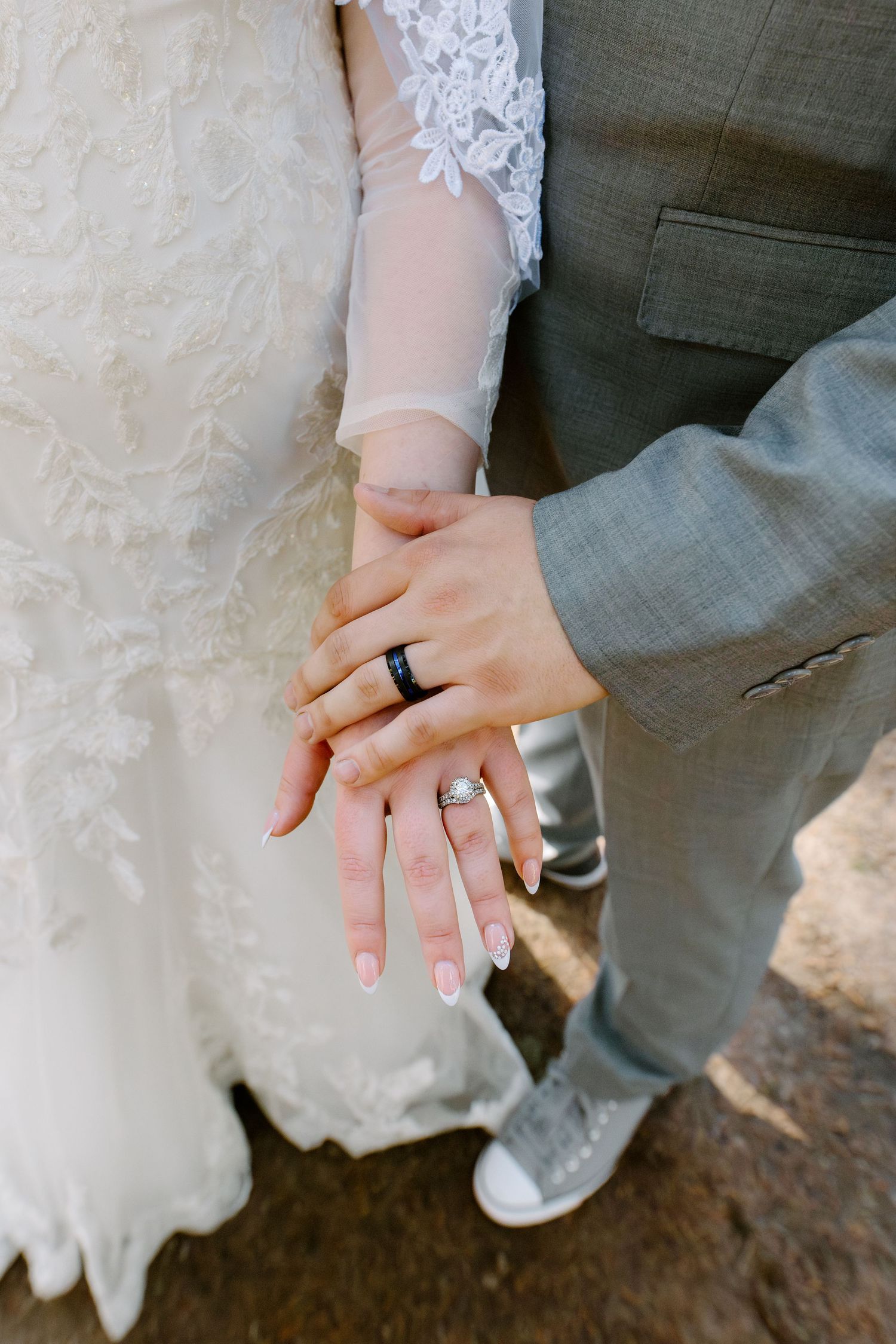 Bride and groom displaying wedding rings, wearing lace gown and gray suit at outdoor ceremony.