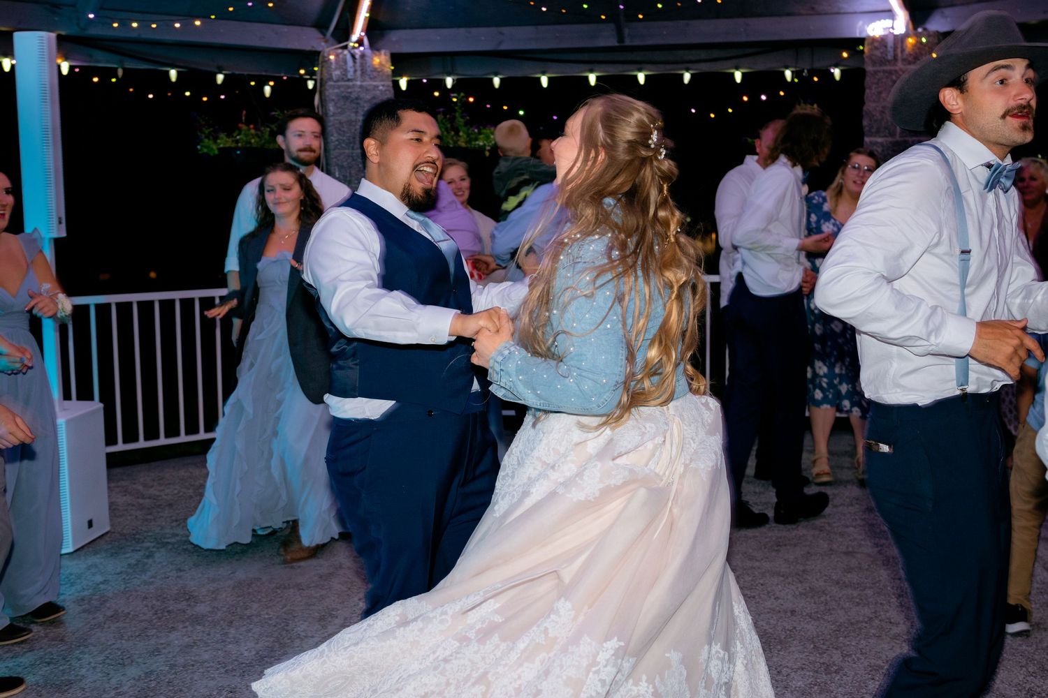 Bride and groom dancing joyfully with guests at a lively outdoor wedding reception under string lights.