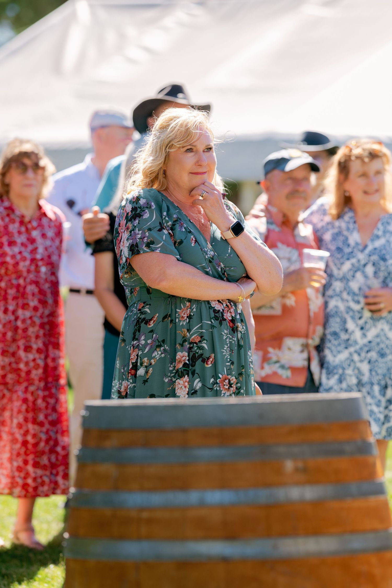 Woman in floral green dress stands thoughtfully by wine barrel at outdoor event.