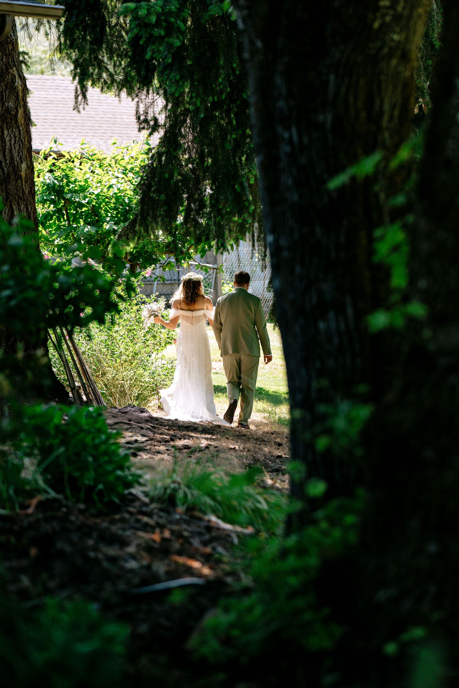 Bride and groom walk hand in hand through lush green wooded path after wedding ceremony.
