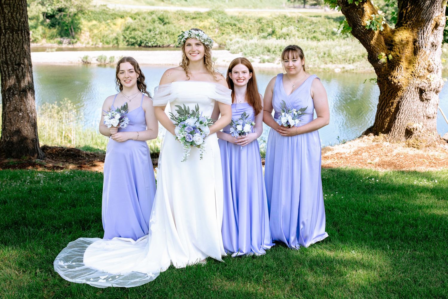 Bride in white gown poses with three bridesmaids in lavender dresses holding bouquets by a pond.
