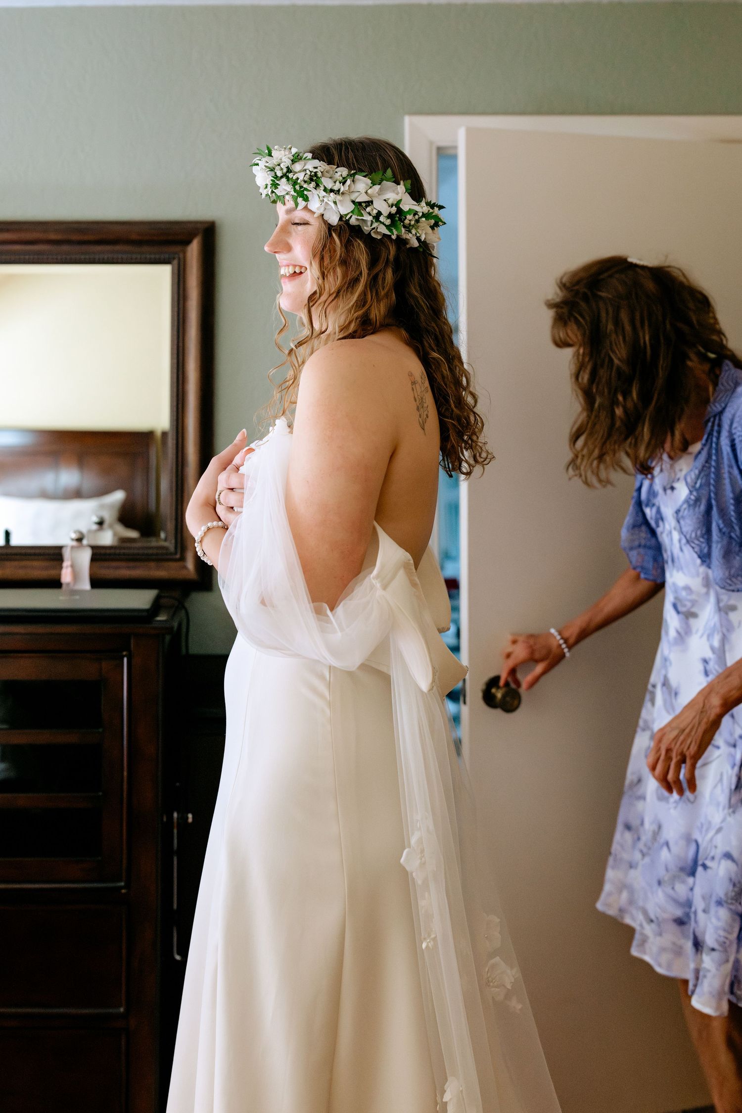 Smiling bride in white off-shoulder gown and floral crown getting ready with help from a woman in blue dress.