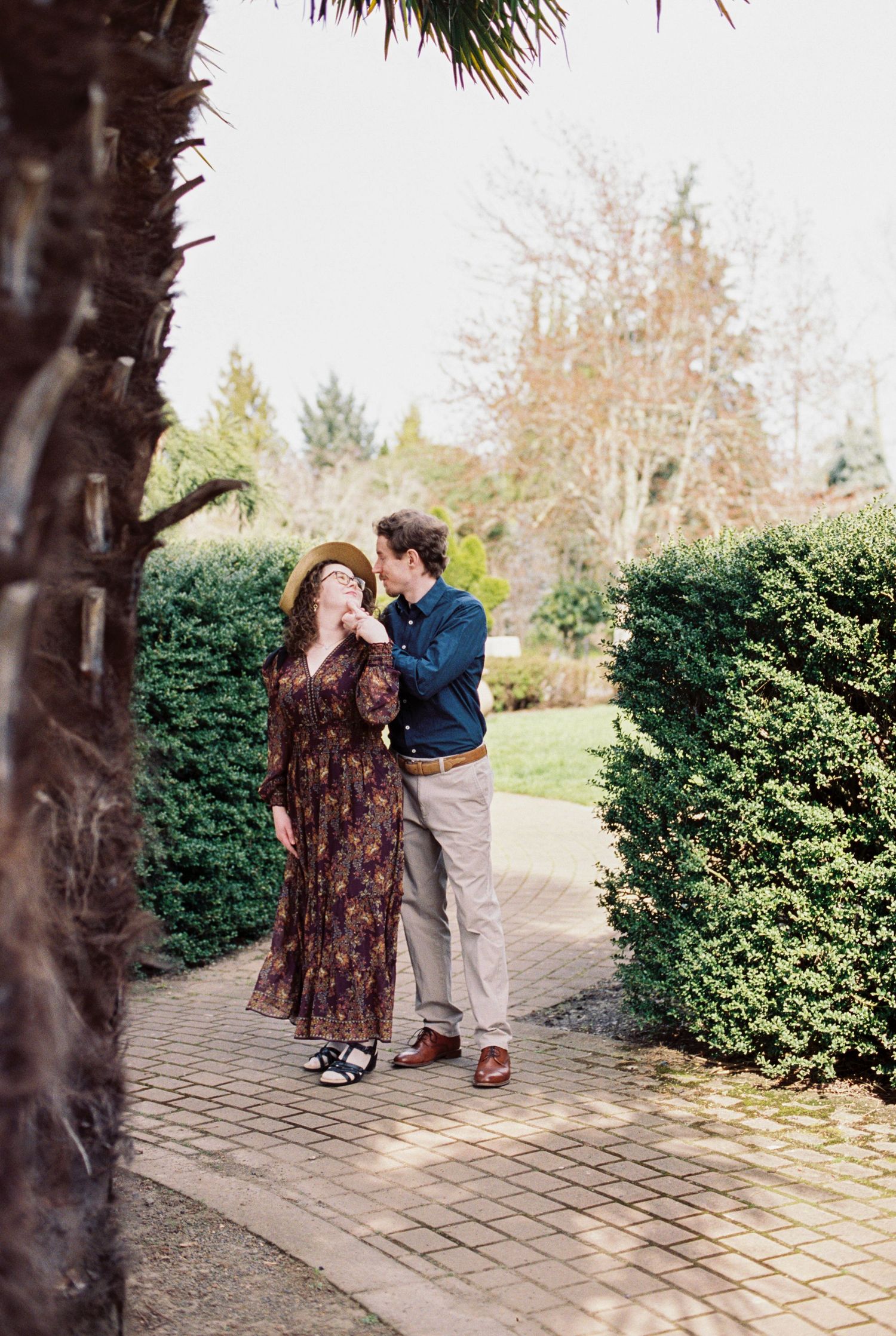 A couple shares intimate moments along a brick pathway surrounded by manicured hedges.