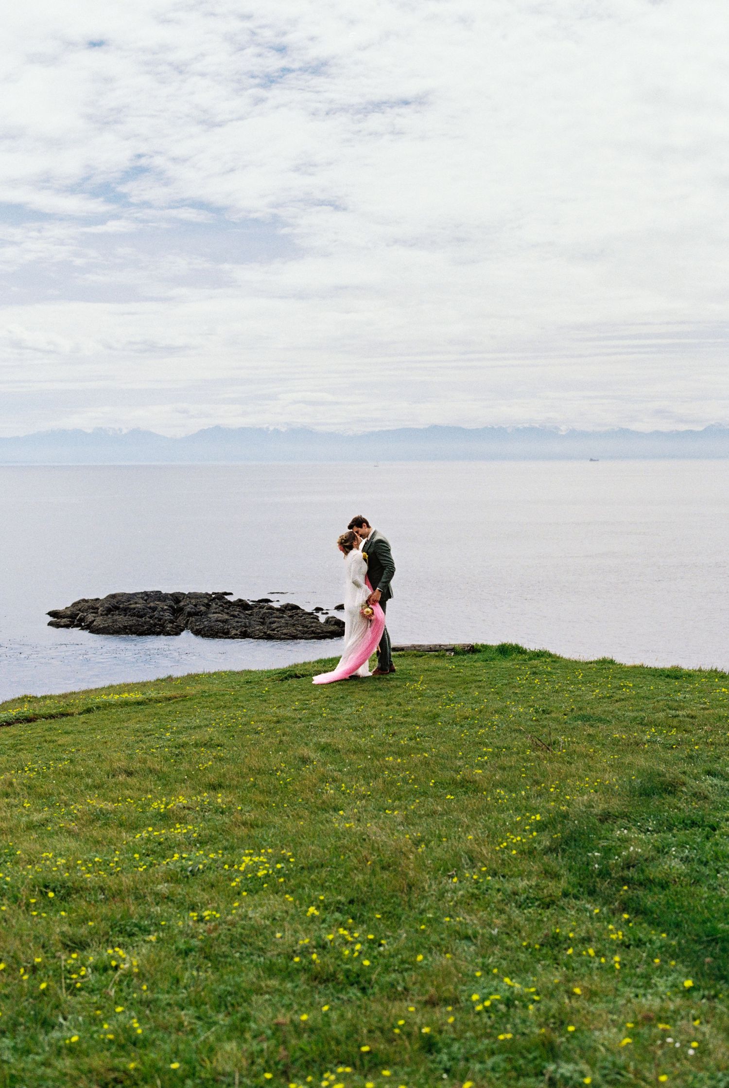 Couple embracing on a lush green cliff overlooking the ocean, bride in pink gown, romantic coastal wedding photo.