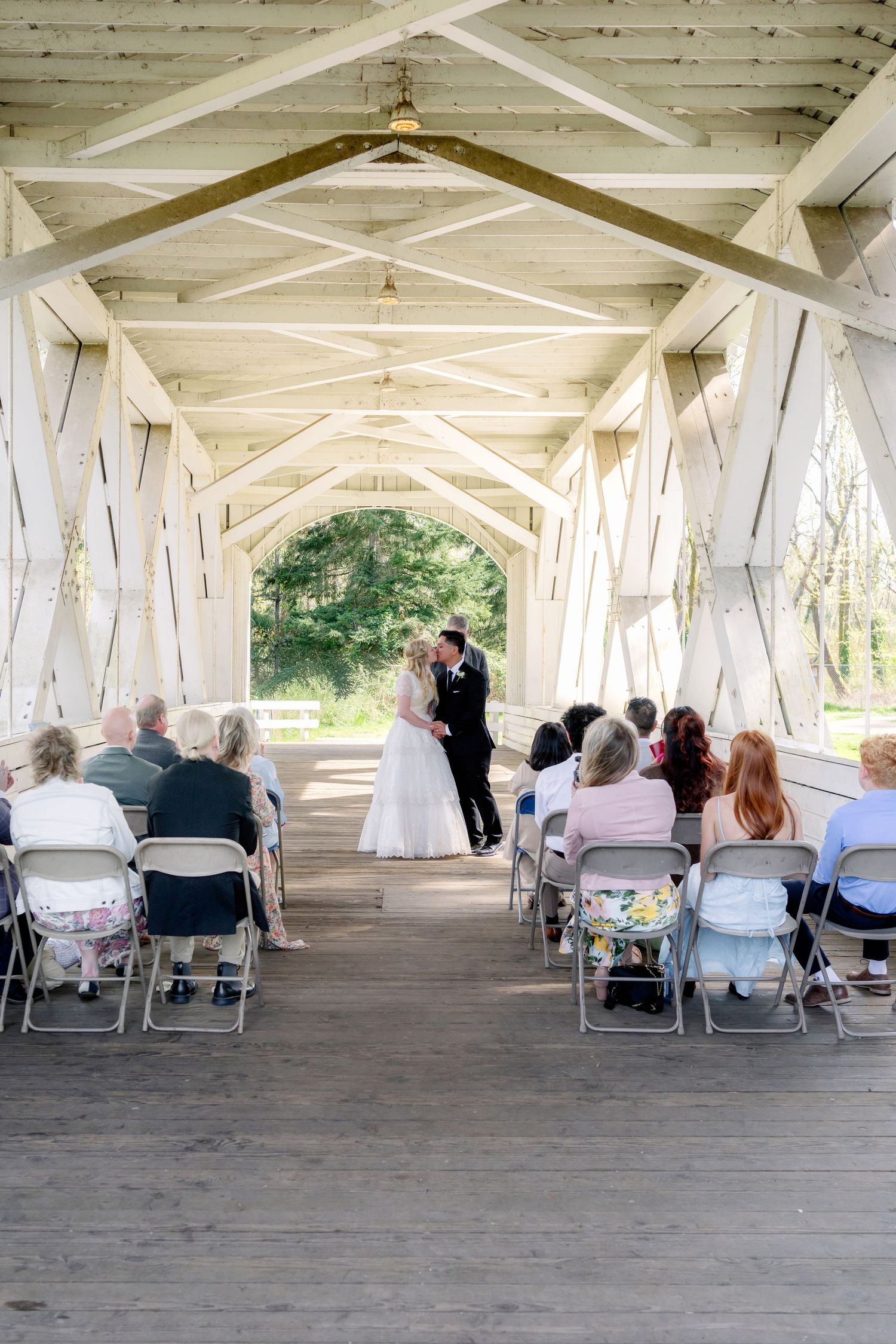 Bride and groom exchanging vows inside a white wooden covered bridge with seated guests.