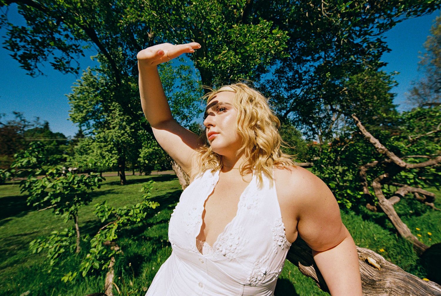 Blonde woman in white halter dress shielding her eyes from the sun in a sunny green park.