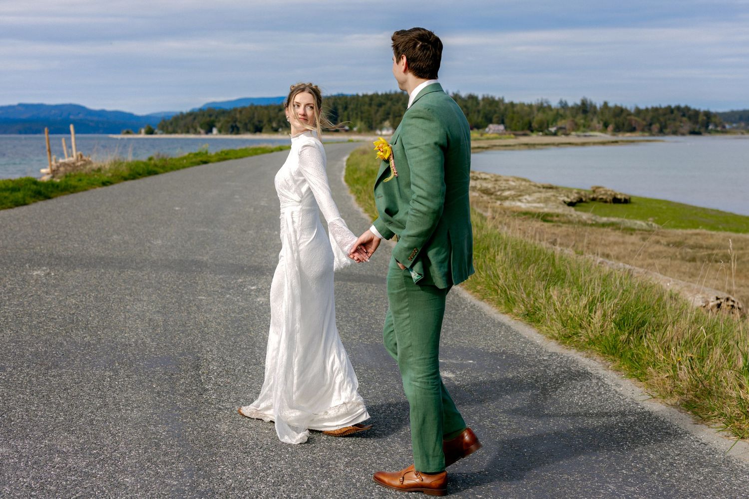 Bride in white gown and groom in green suit holding hands on a scenic coastal road with water views.