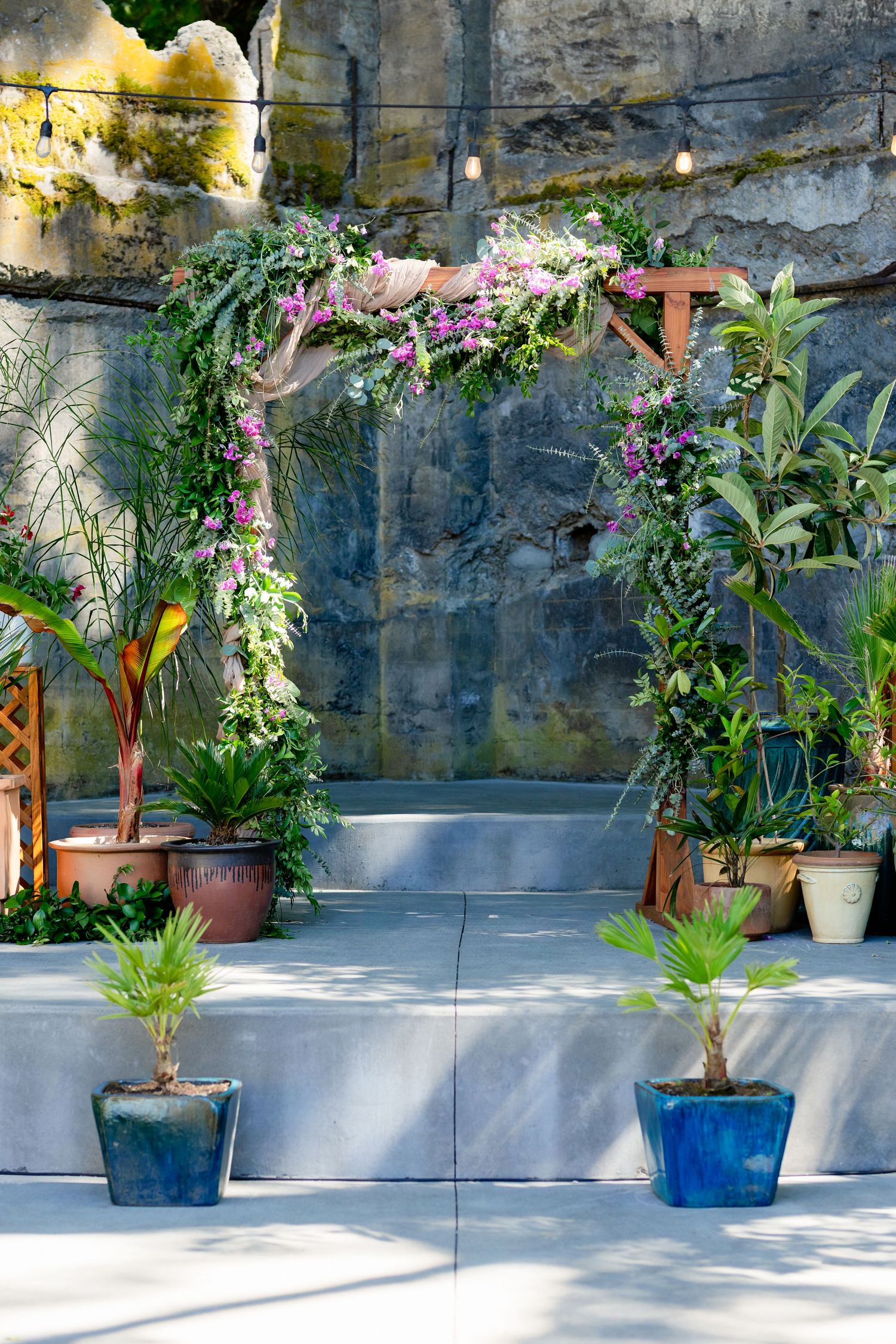 Floral wedding arch adorned with pink flowers and greenery surrounded by potted tropical plants on a concrete stage.