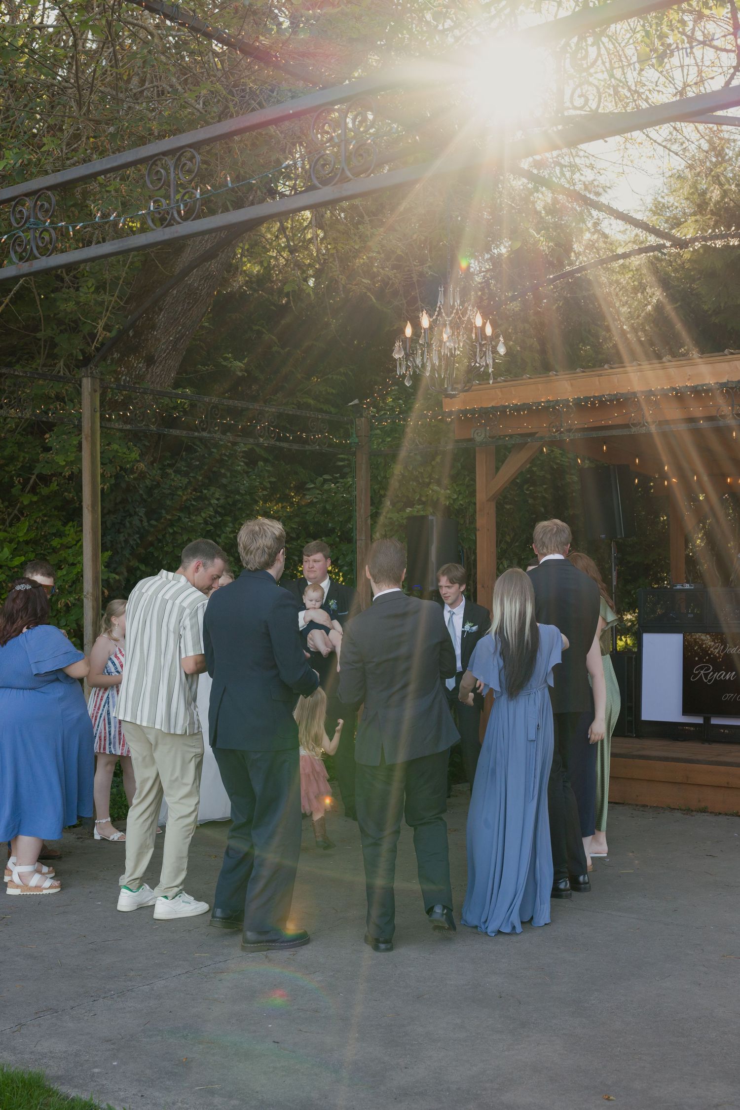 Wedding guests mingle on outdoor patio under pergola with chandelier as golden sunlight streams through the trees.