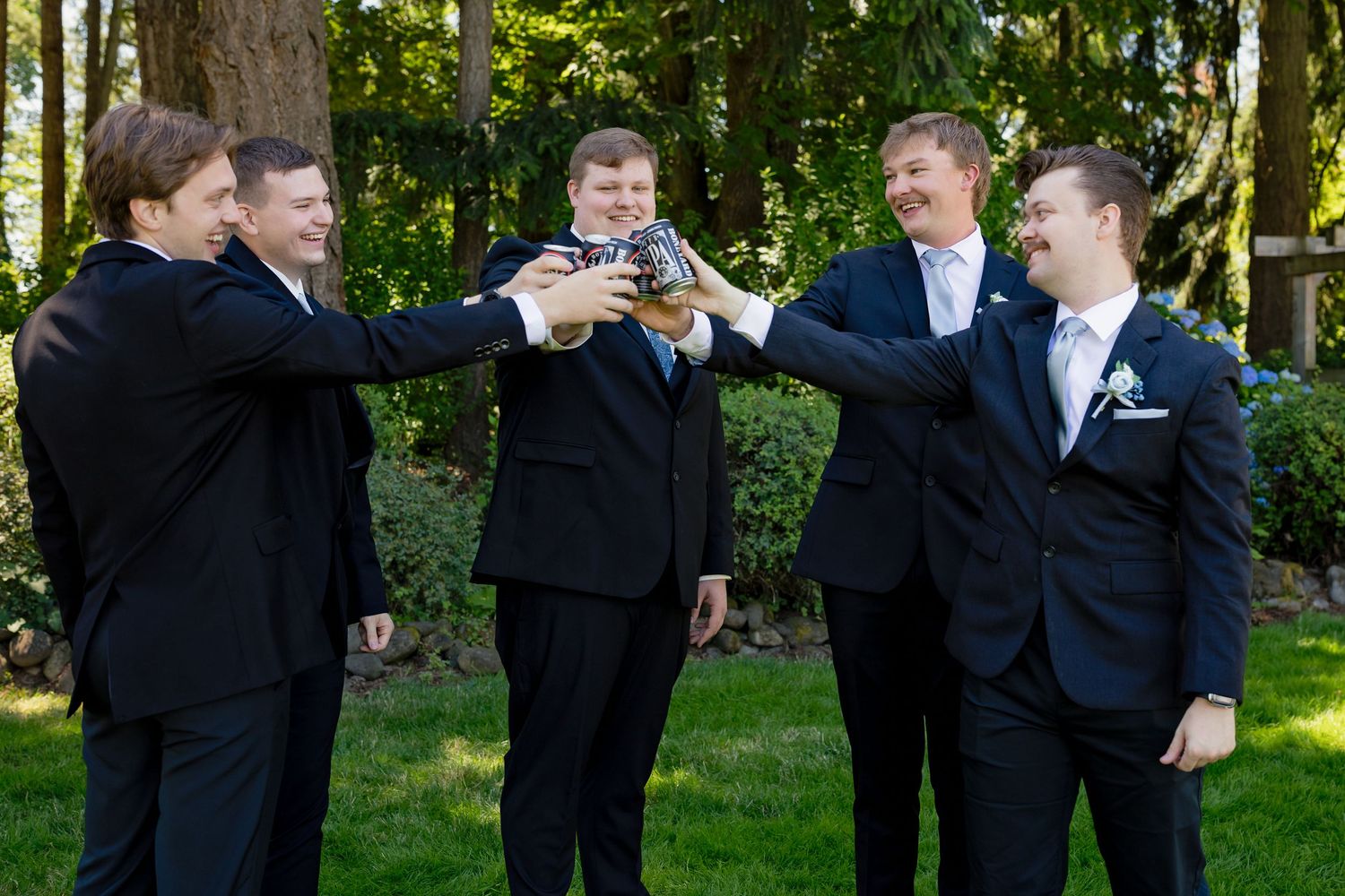 Five groomsmen in black suits toasting with glasses outdoors at a wedding celebration among green trees.