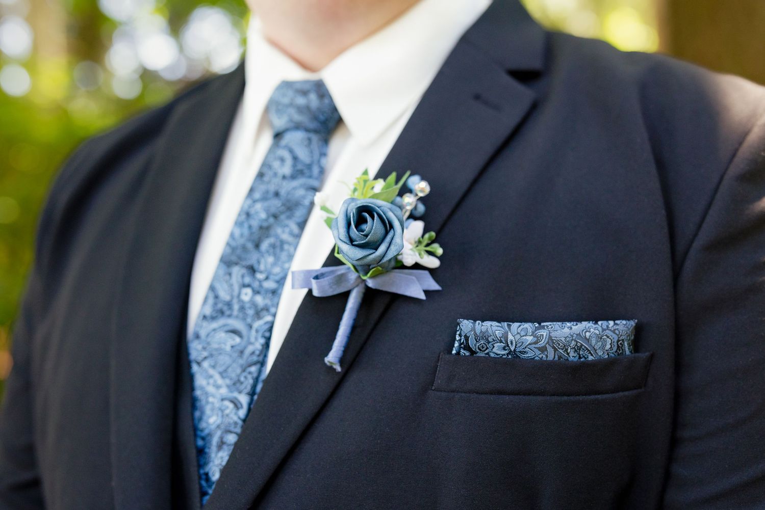Groom wearing dark suit with blue paisley tie, matching pocket square, and elegant navy rose boutonniere.