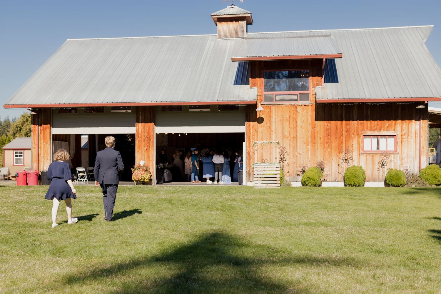 Guests walk toward a large rustic wooden barn wedding reception venue on a sunny afternoon.