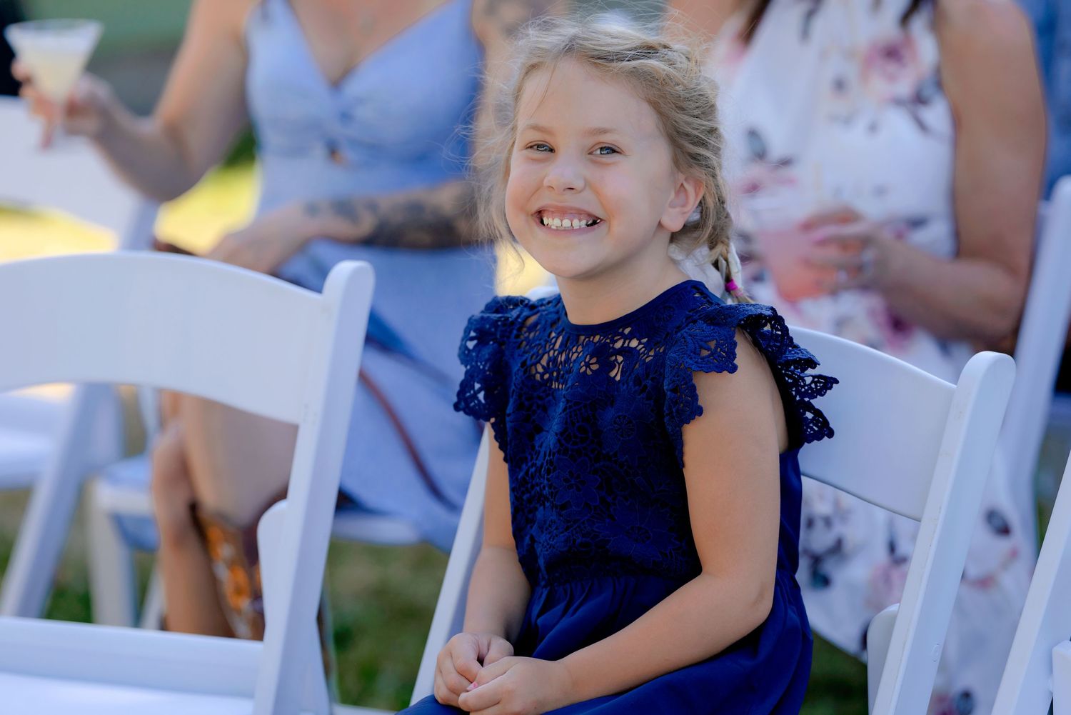 A young girl in a navy blue lace dress smiles joyfully while seated at an outdoor wedding ceremony.
