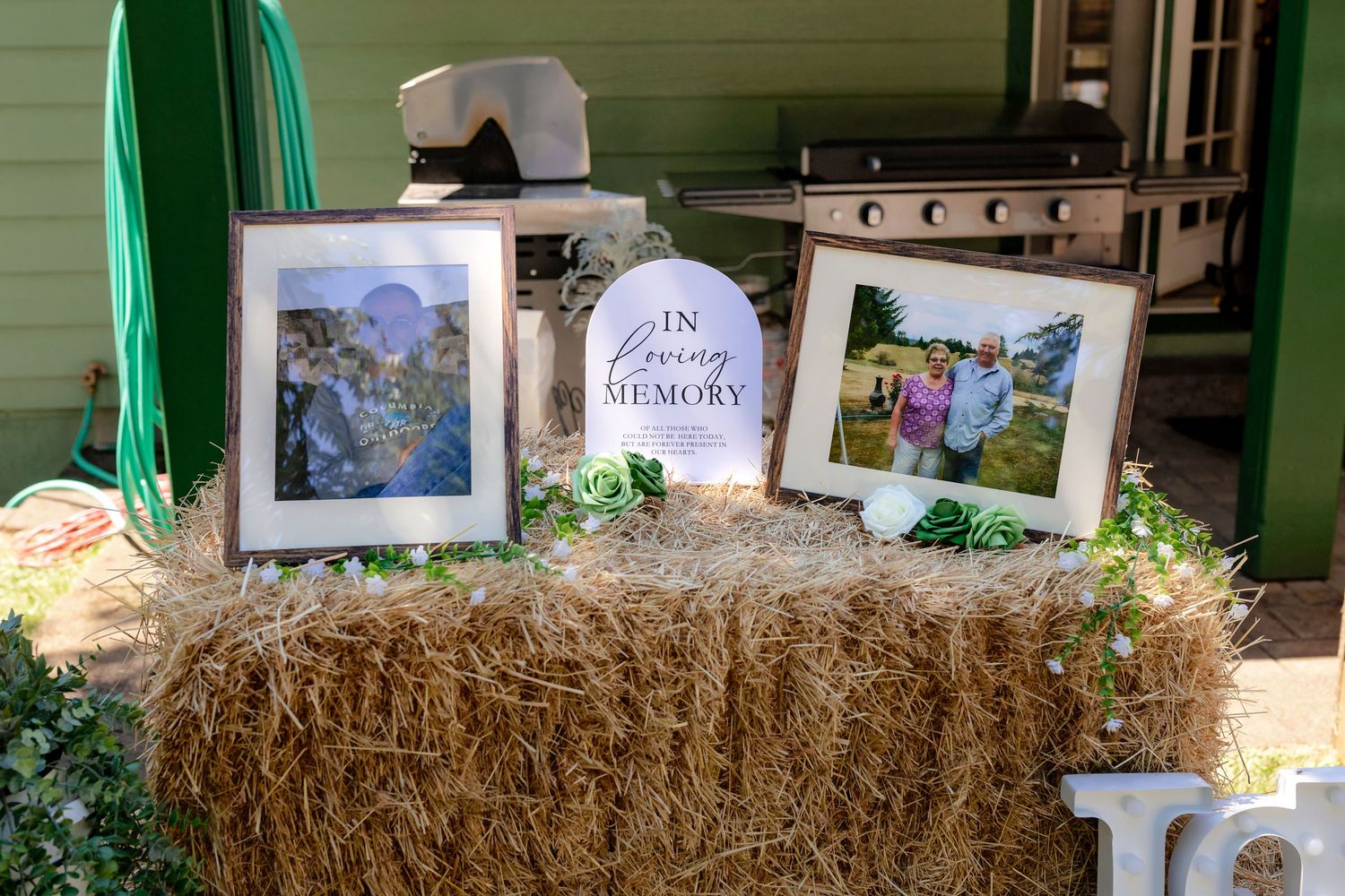 A rustic hay bale memorial display with framed photos and an In Loving Memory sign at an outdoor wedding.