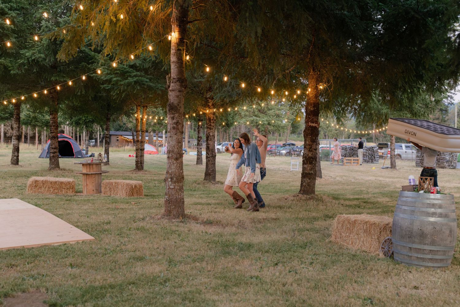 Guests dancing under string lights among trees with hay bales at outdoor wedding.