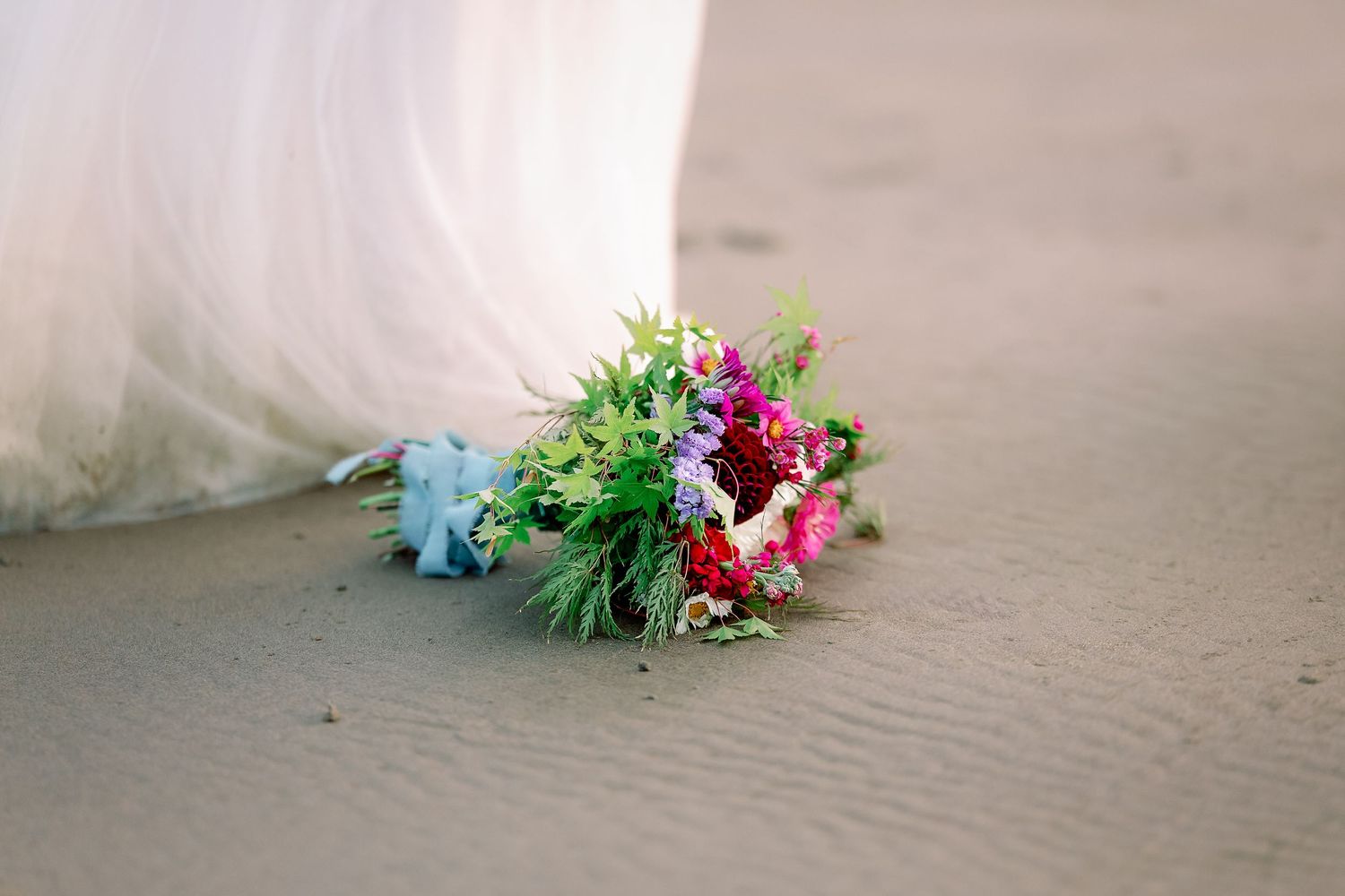 A white wedding dress train and colorful floral bouquet lying on a sandy beach during sunset.