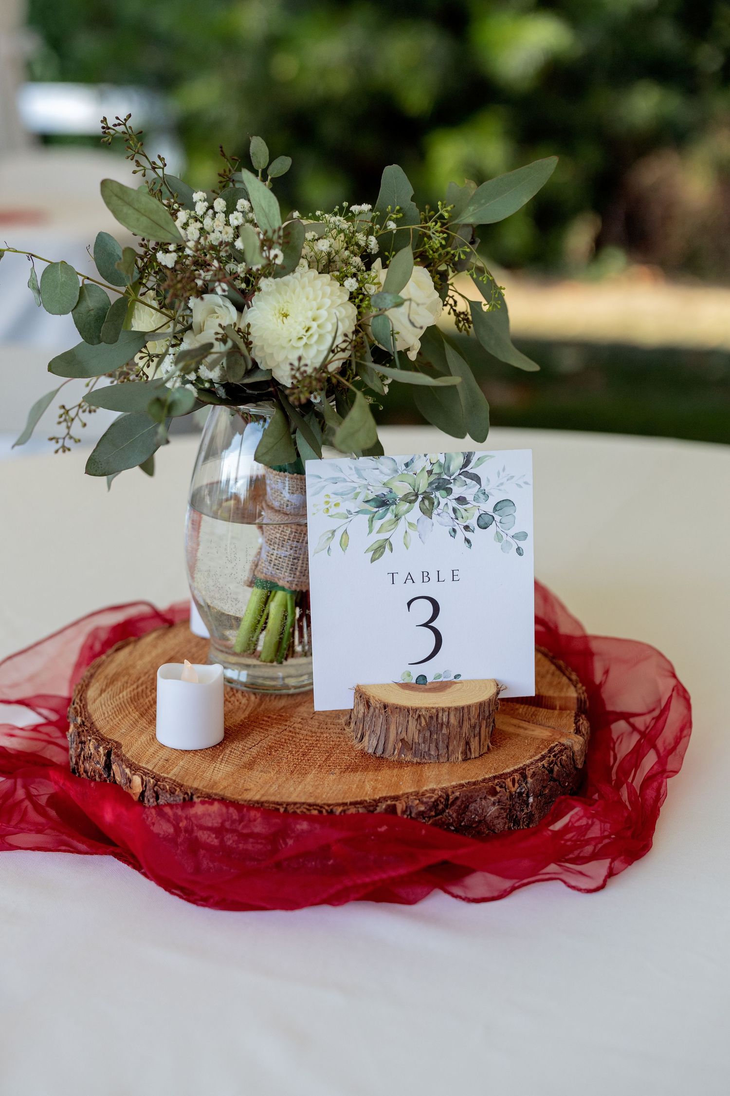 Rustic wedding table centerpiece with eucalyptus on wood slice, red chiffon runner, and table number card.
