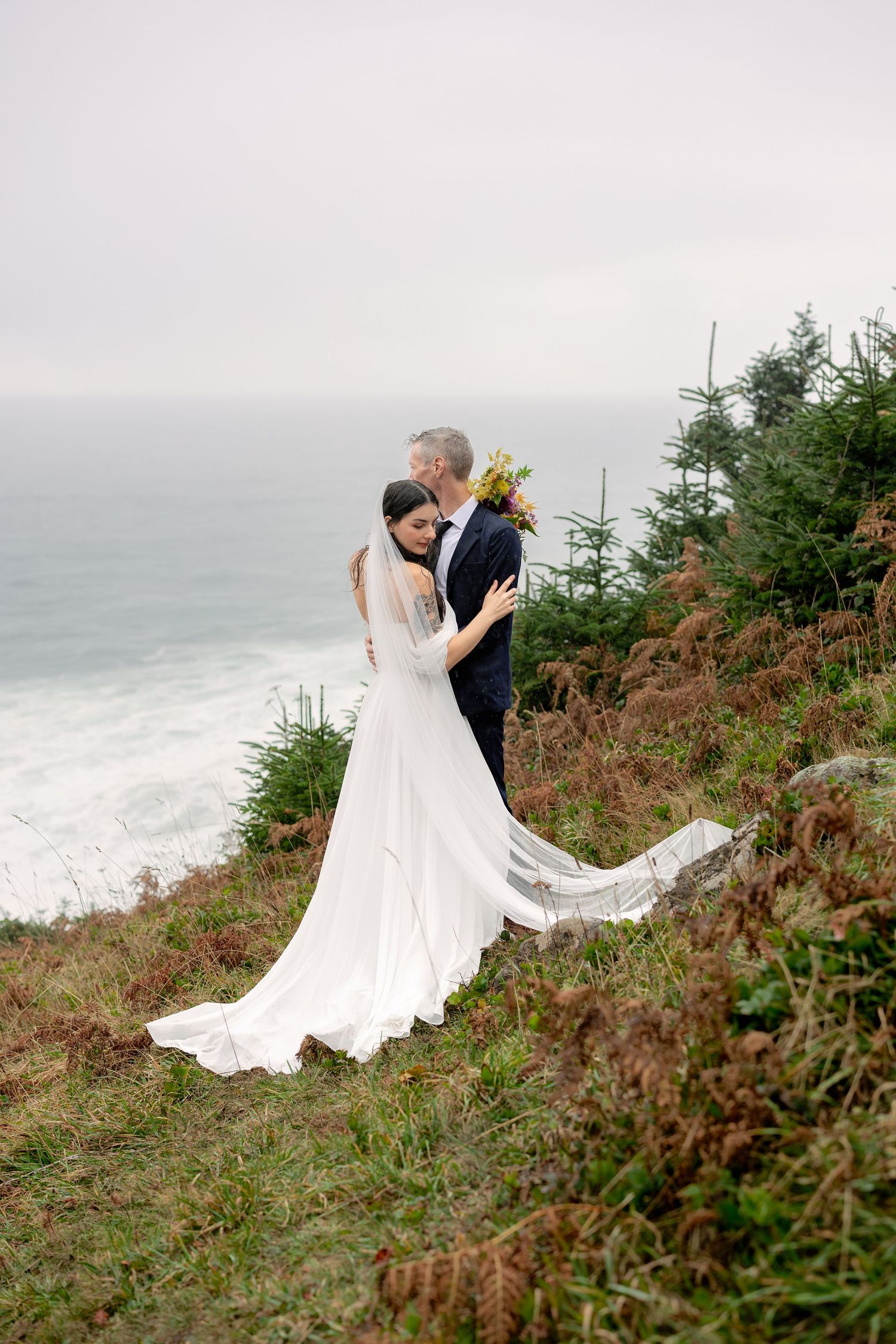 A couple embraces on a misty coastal cliff with a flowing white wedding dress and dramatic ocean backdrop.