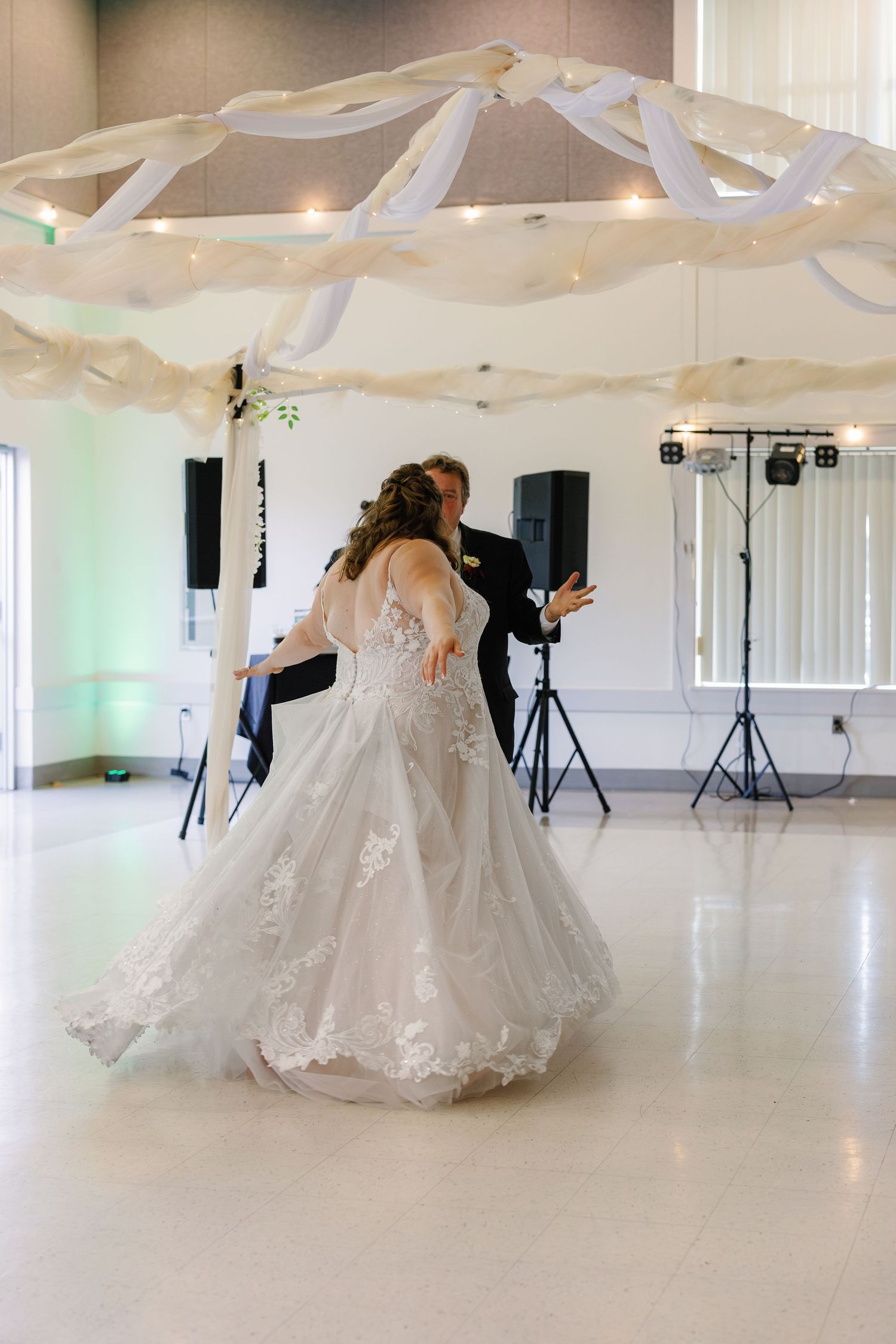 White wedding dress twirls gracefully across a bright indoor dance floor with draped fabric decorations.