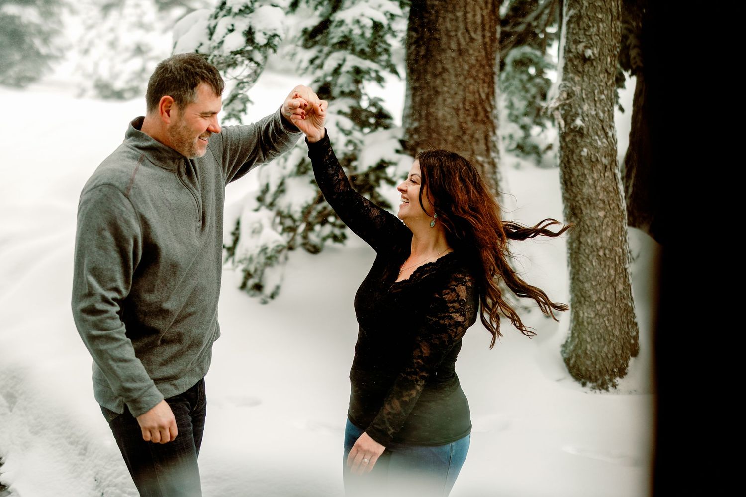 A couple dances together in the snow surrounded by winter trees, sharing a joyful moment outdoors.
