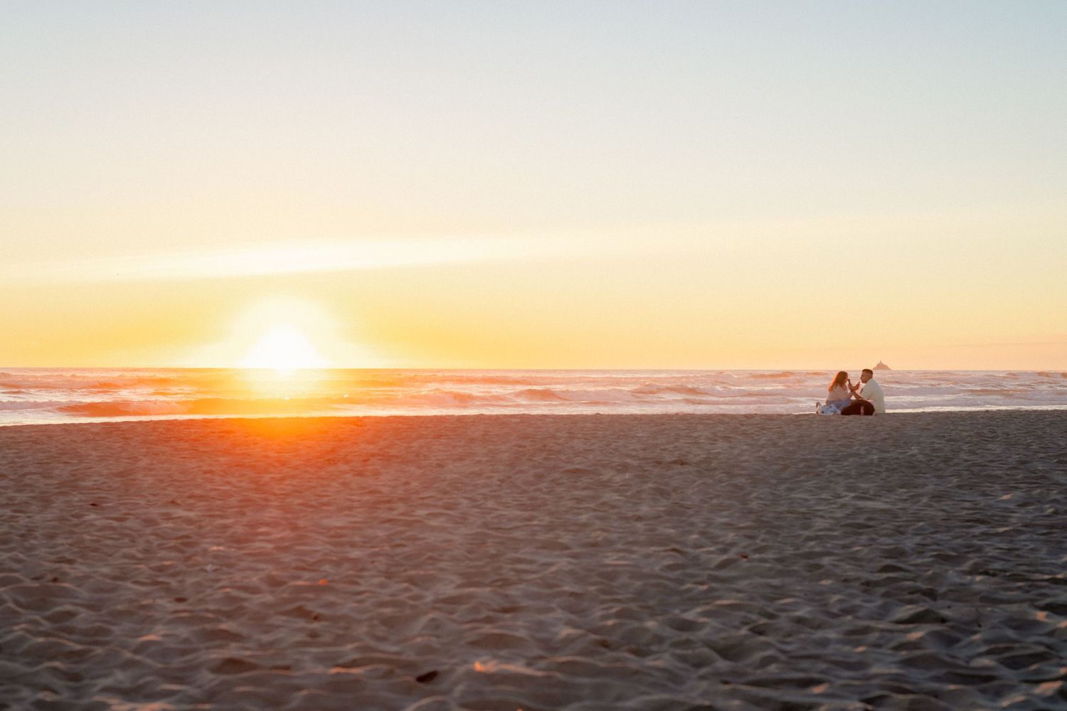 Couple sitting on a sandy beach watching a golden sunset over the ocean horizon.